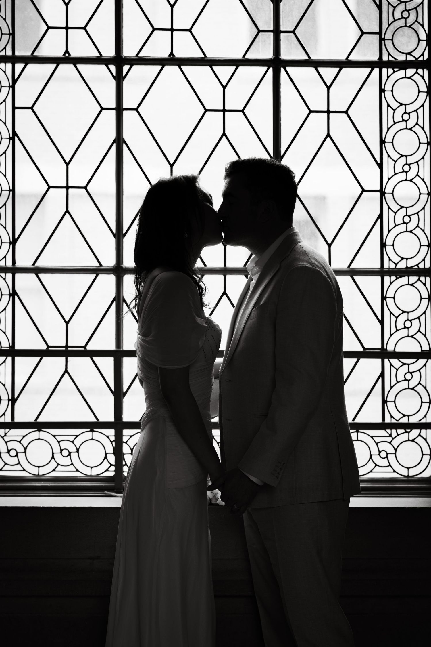 Silhouette of a couple kissing while holding hands in front of an ornate stained glass window in black and white.