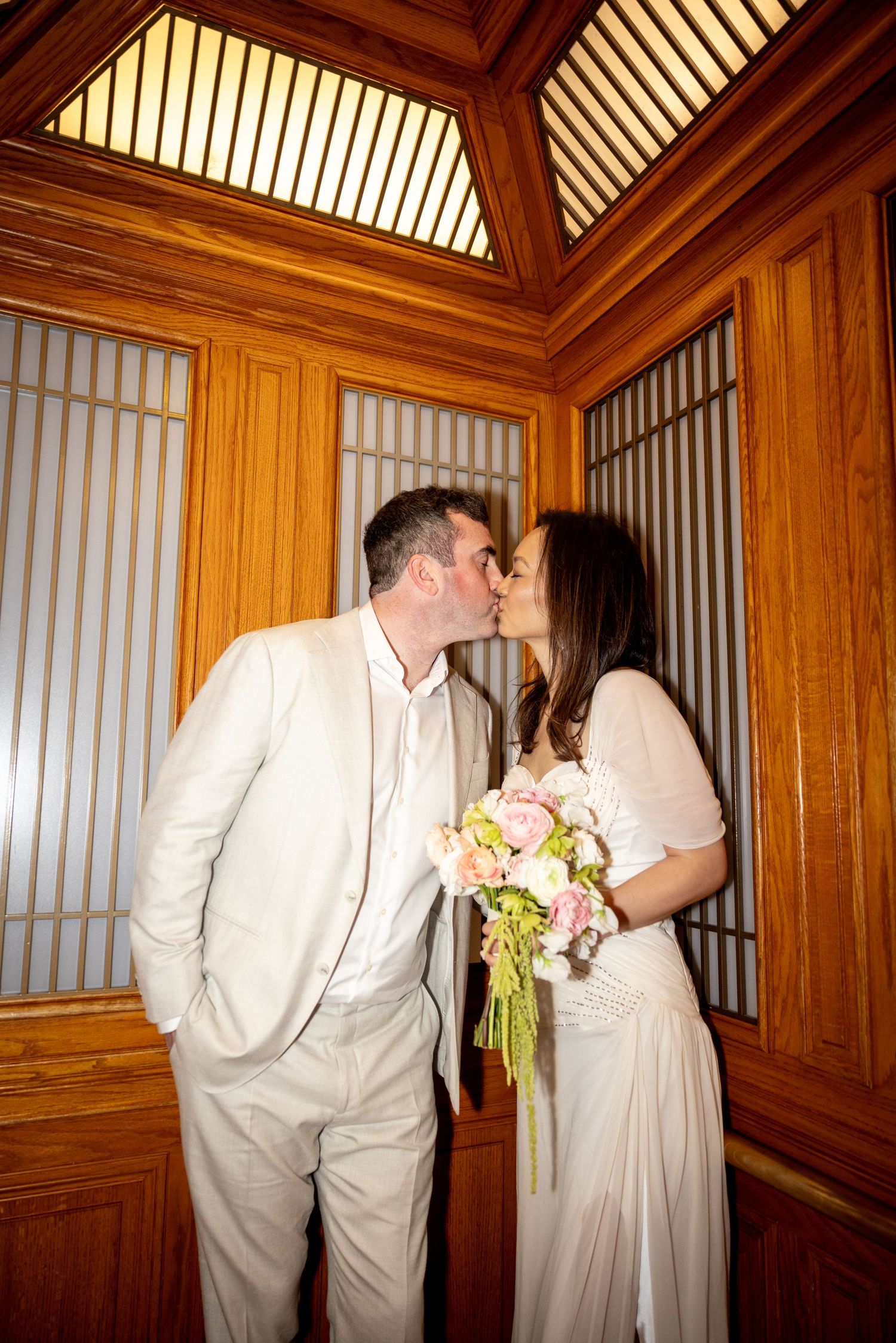 Bride and groom sharing a kiss in a wood-paneled room, bride holding a pink and white bouquet.