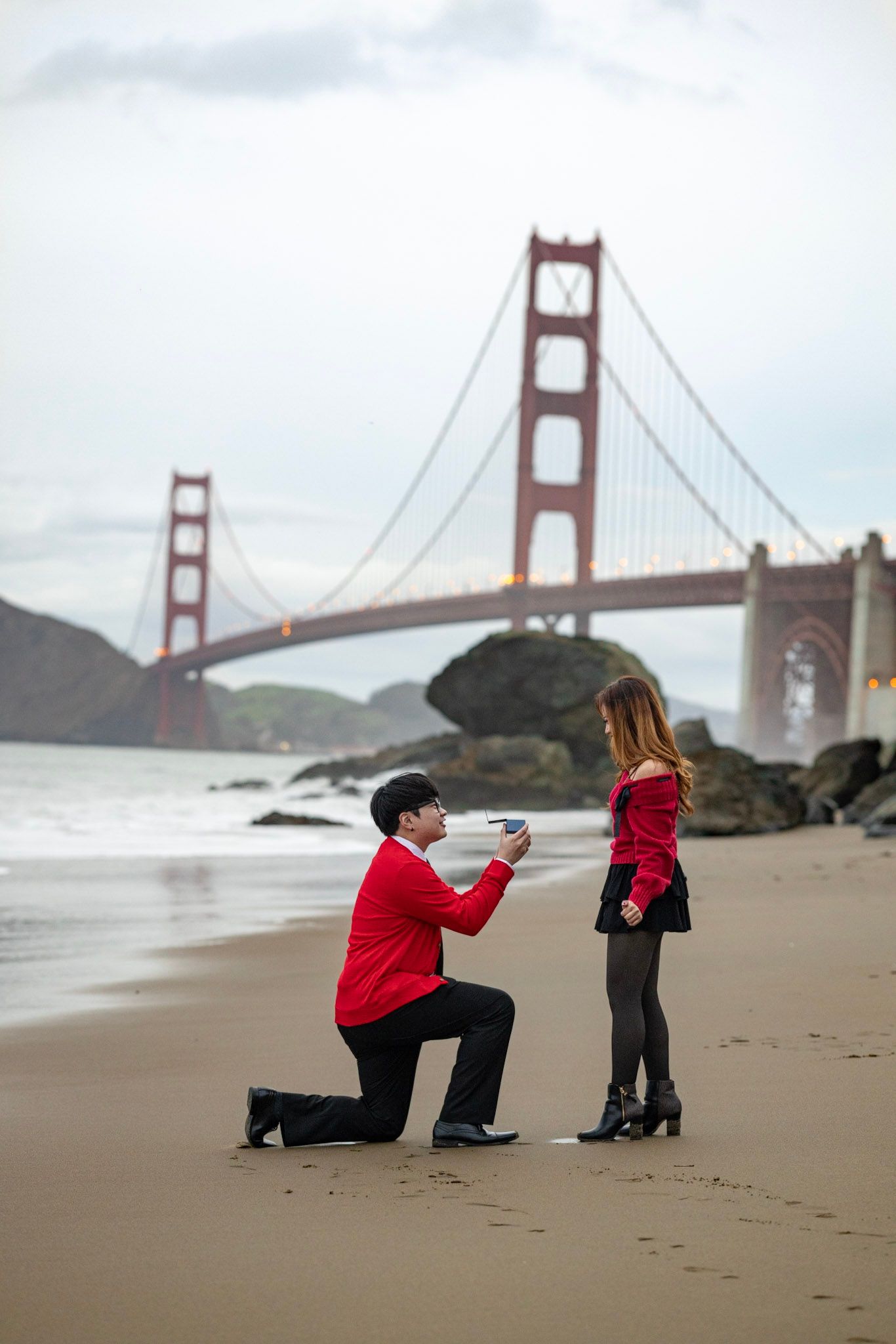 A romantic proposal scene unfolds on Baker Beach with the iconic Golden Gate Bridge in the background.