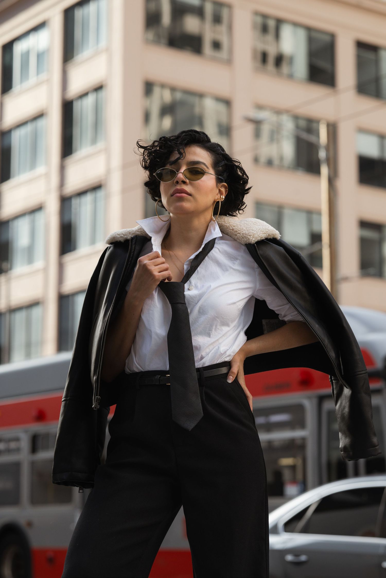 Stylish woman in business attire poses confidently against urban backdrop with sunglasses and black blazer.
