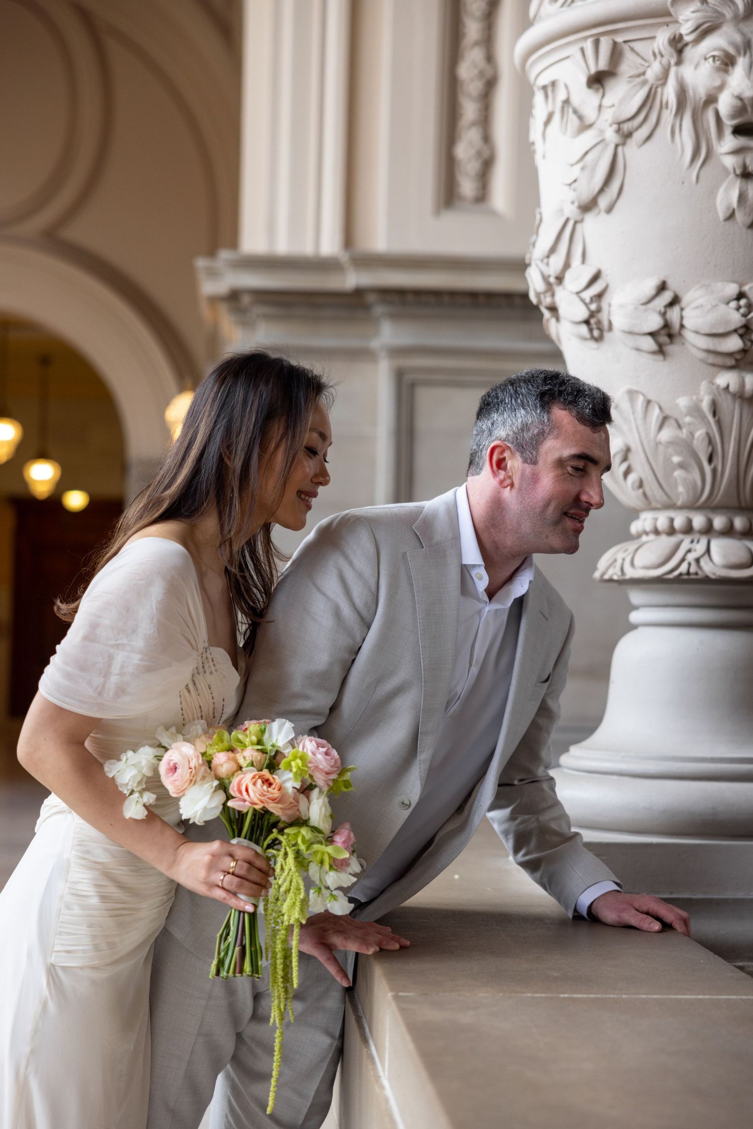 A bride holding a pastel bouquet and groom in a gray suit share a joyful moment beside an ornate column.