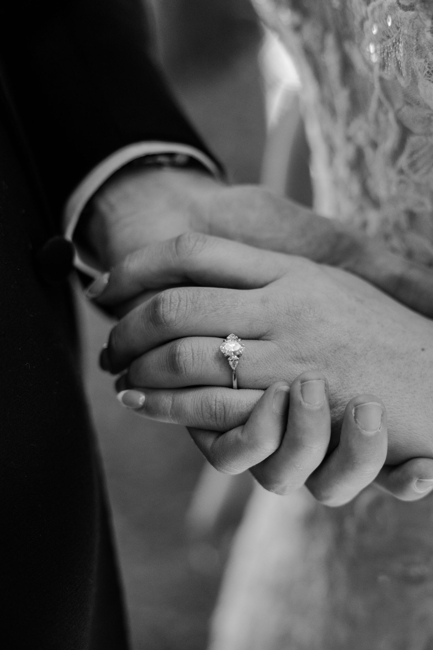 Black and white photo of a bride and groom holding hands, showcasing a diamond engagement ring on the bride's finger.
