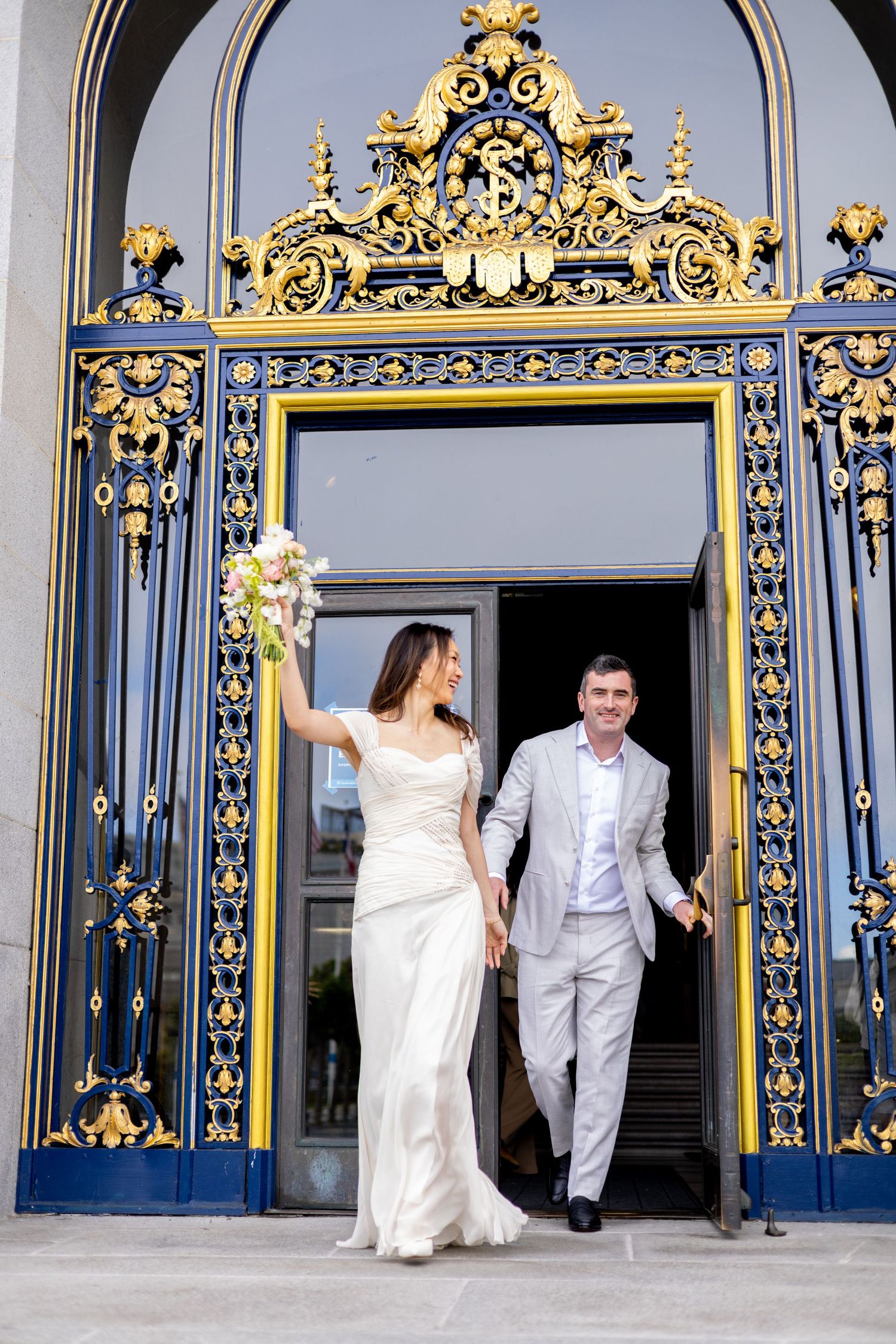 Bride and groom exit ornate gold and blue doors of San Francisco City Hall holding hands after wedding ceremony.