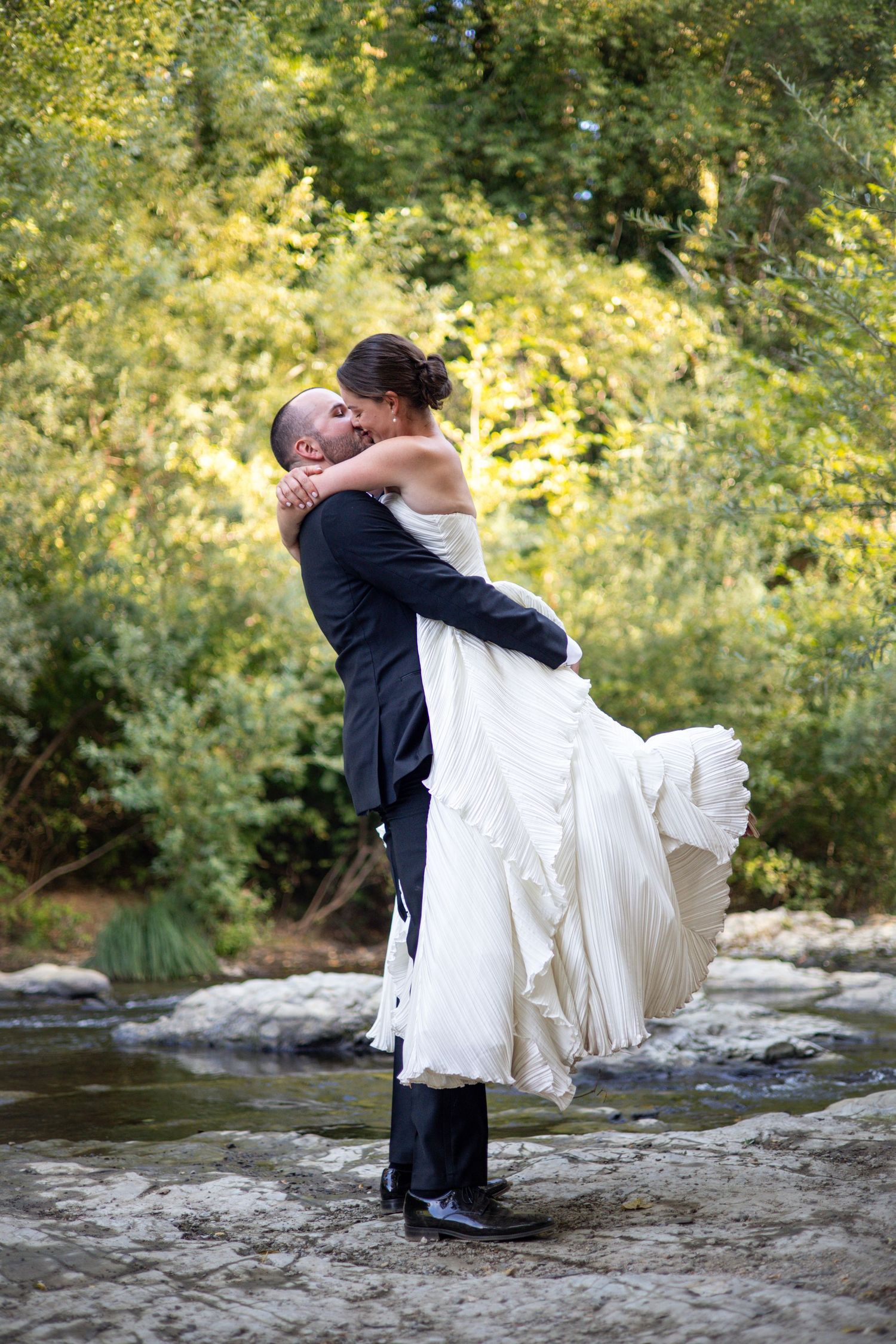 Groom lifting bride in white dress, sharing a romantic kiss outdoors by a stream with lush green trees.