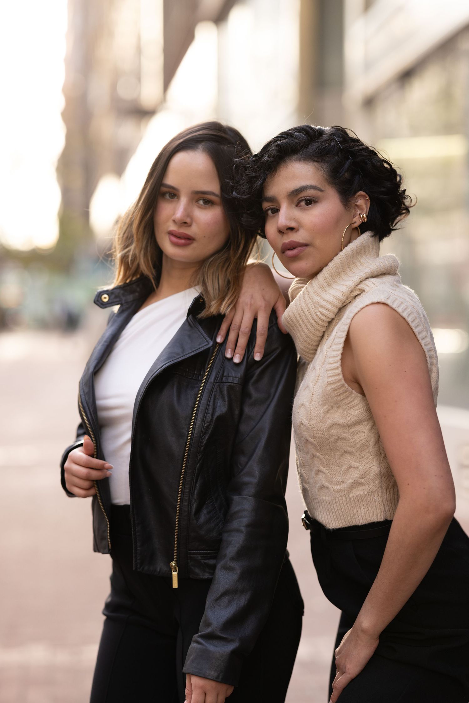Two people pose together in black and beige outfits for a fashion photoshoot on an urban street.