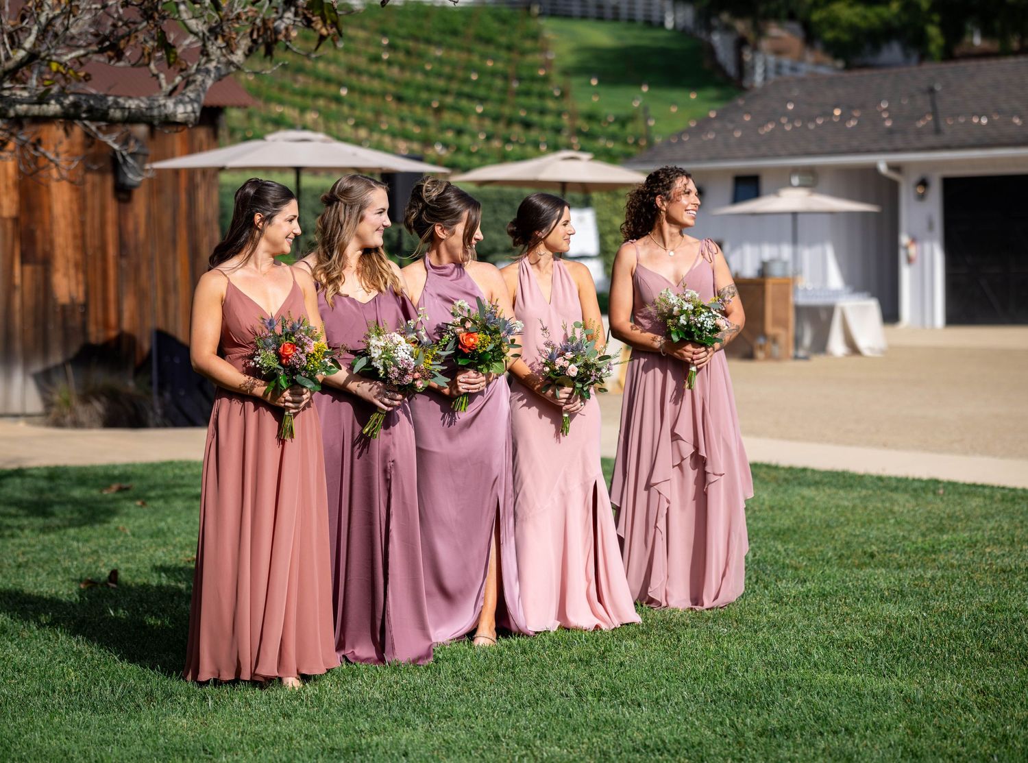 Four bridesmaids in pink dresses holding colorful bouquets walk across green lawn at outdoor wedding venue.