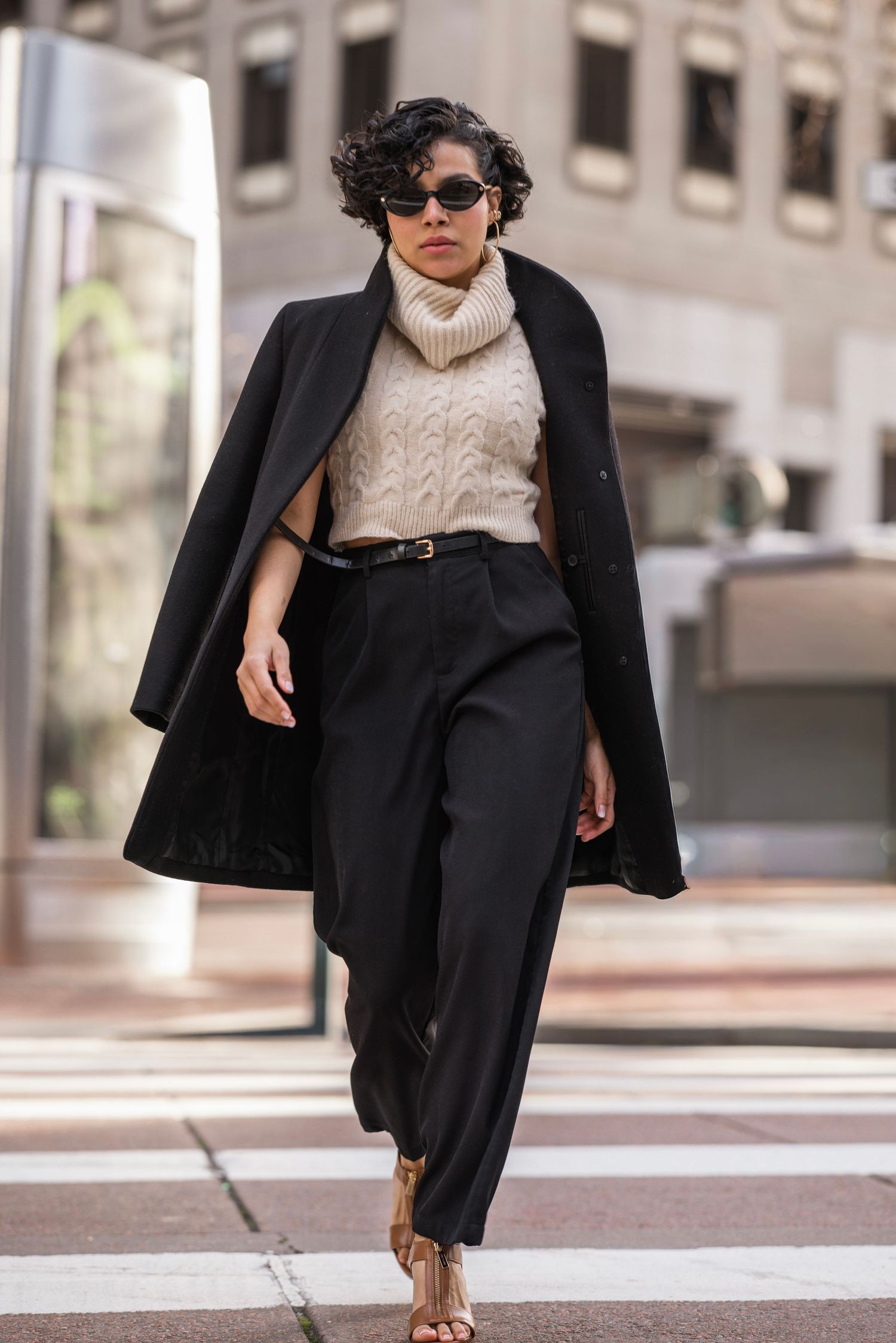 Fashion blogger wearing cream turtleneck sweater, black blazer, trousers and sunglasses poses in urban setting.