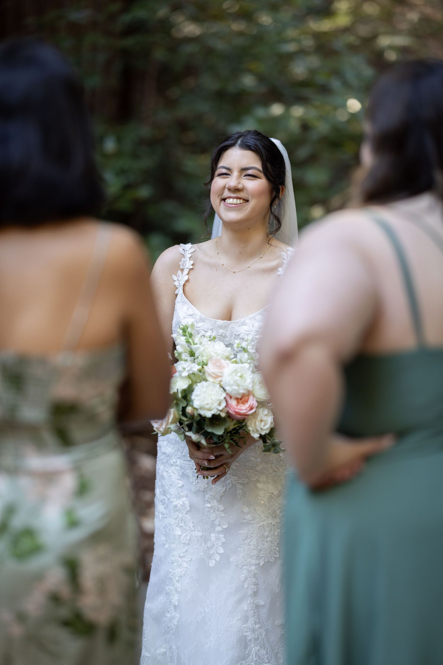 Smiling bride in lace gown holds pink and white bouquet, flanked by bridesmaids in green dresses outdoors.