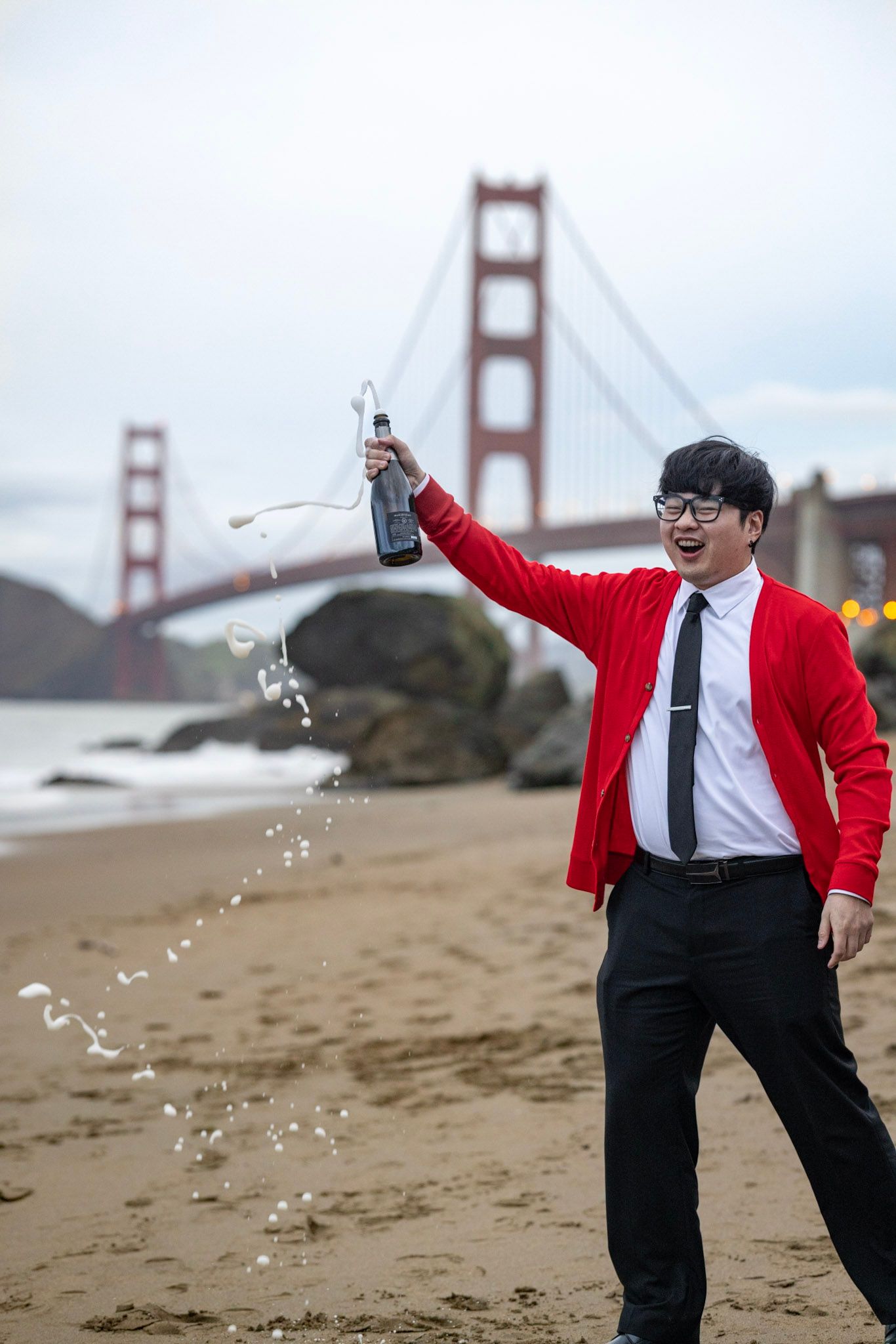 A person in a red shirt and tie throws water on a beach with the Golden Gate Bridge visible in the background.