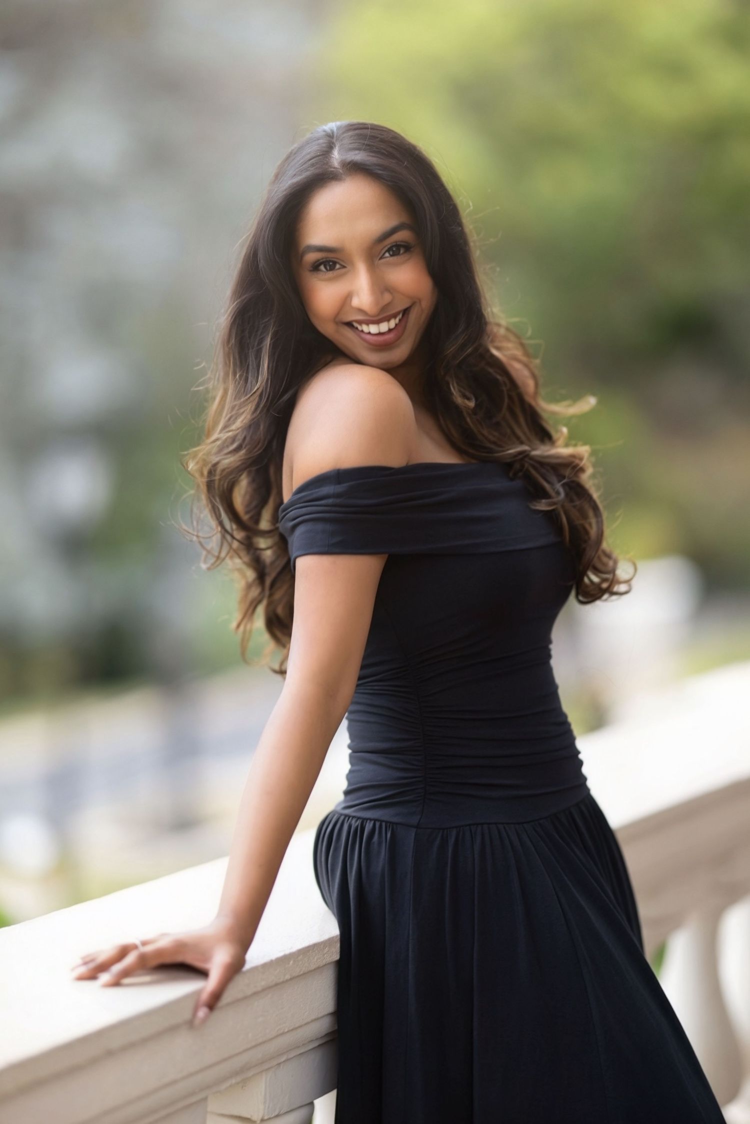 Woman in elegant black off-shoulder dress poses outdoors on balcony in natural lighting.