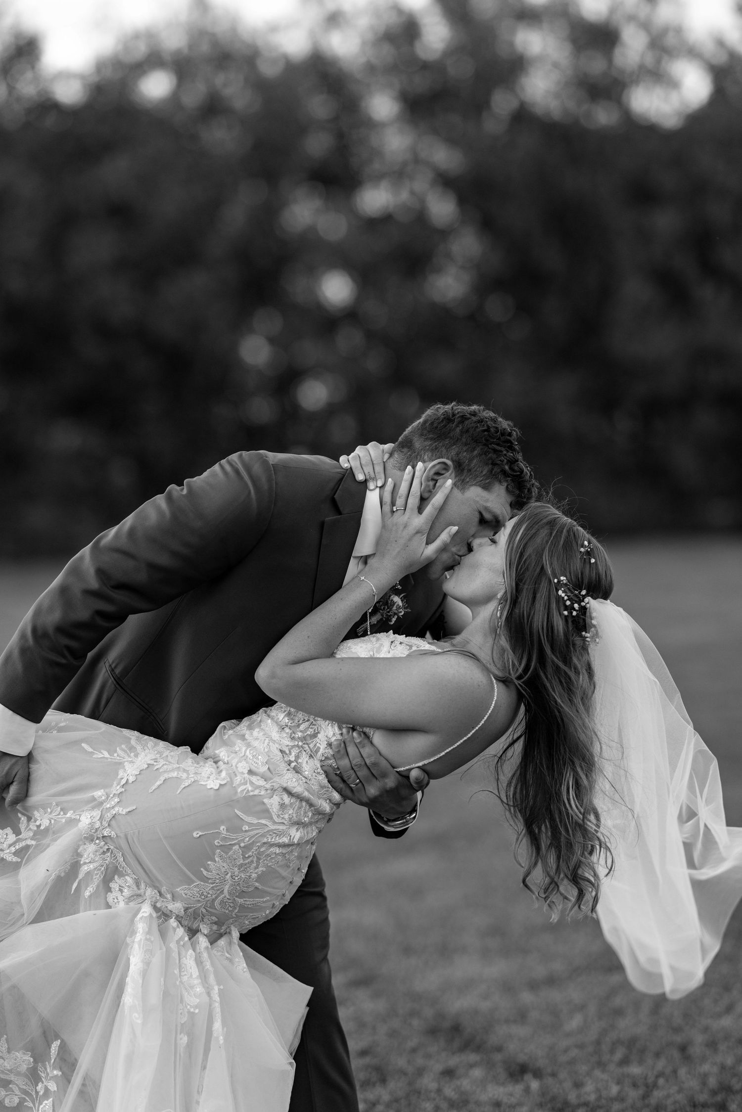 A romantic black and white wedding photo shows a couple sharing a passionate kiss outdoors with the bride in a lace gown and veil.