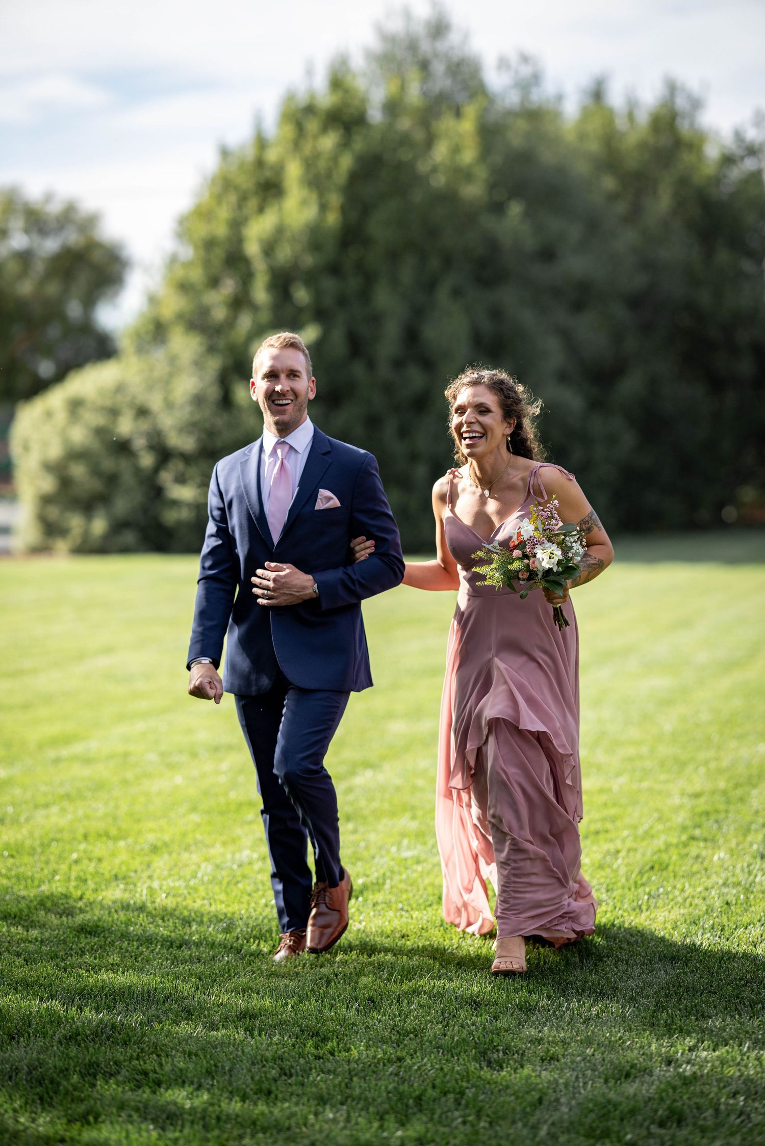 A smiling groomsman in a navy suit escorts a bridesmaid in a pink gown across a lush green lawn.