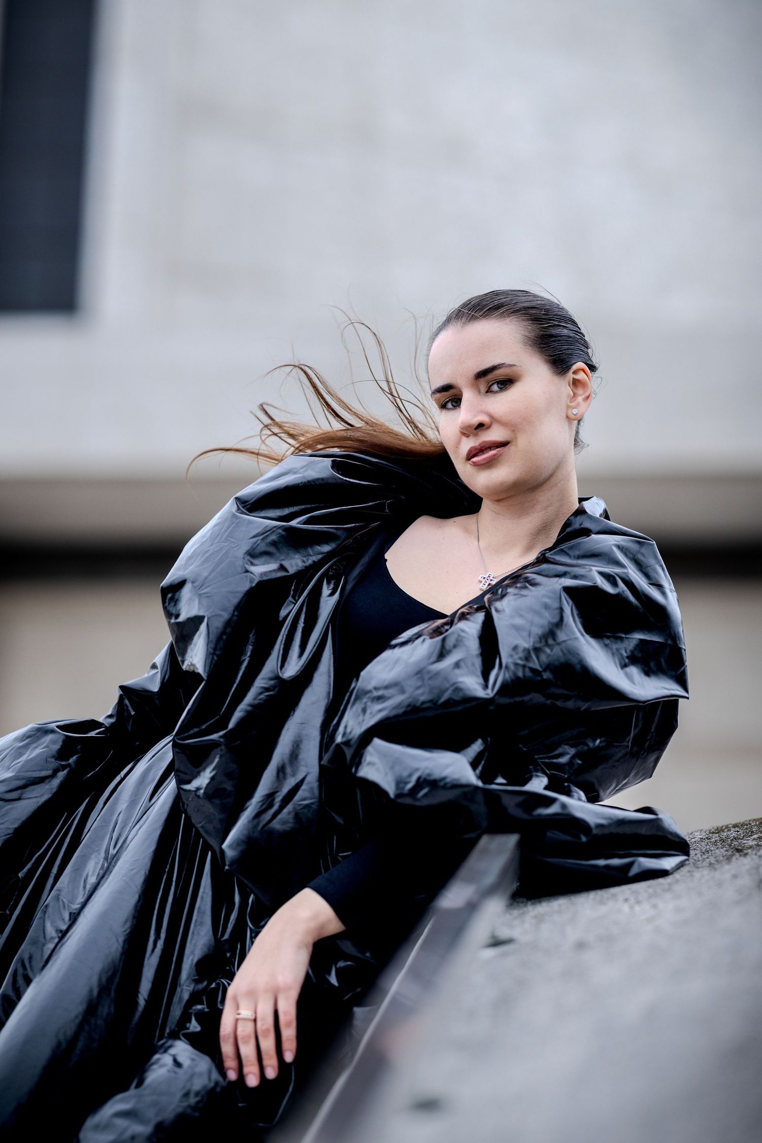 Woman in black shiny coat with wind-blown hair posing outdoors against a concrete building backdrop.