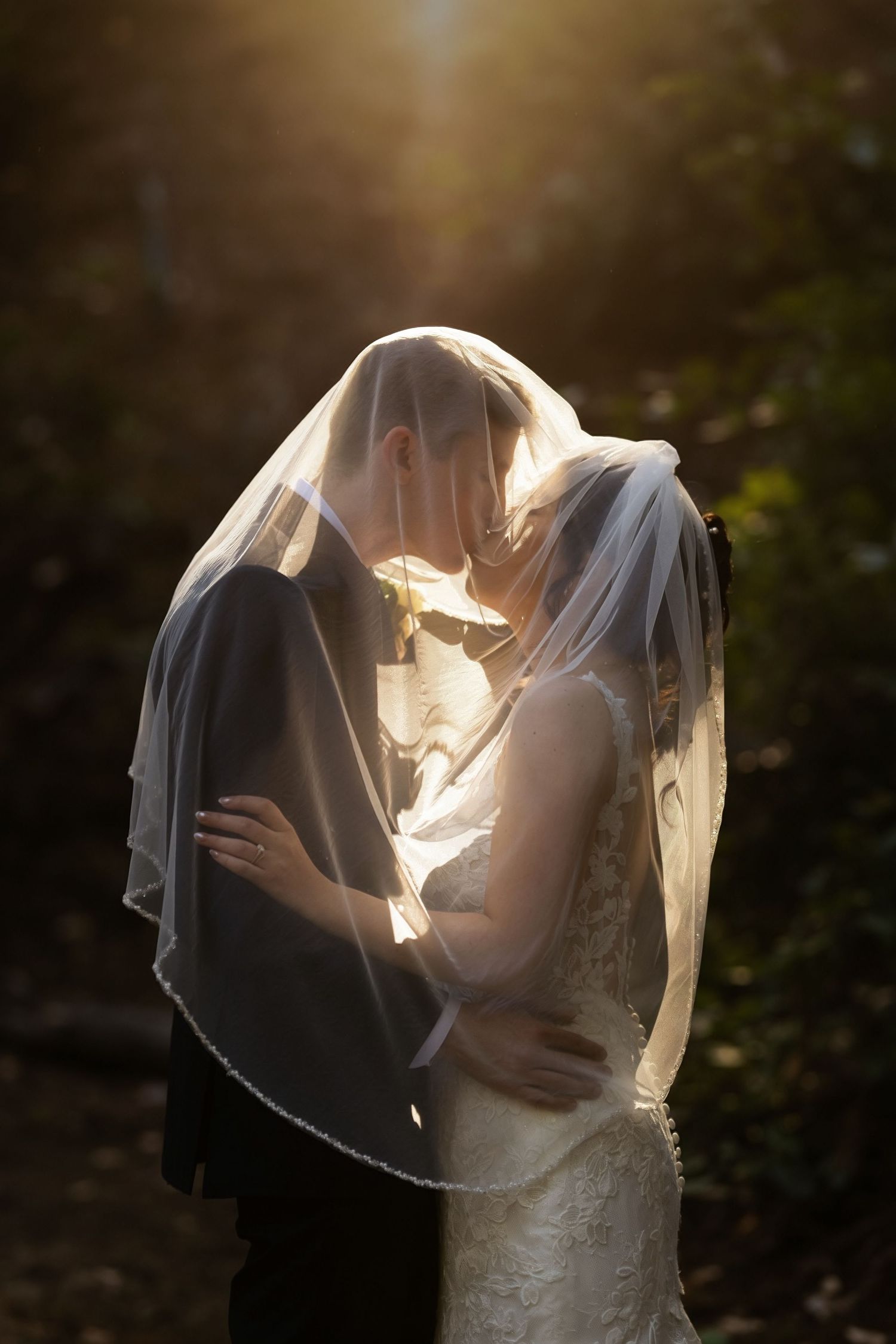Bride and groom kissing under wedding veil with golden backlight in outdoor forest setting.