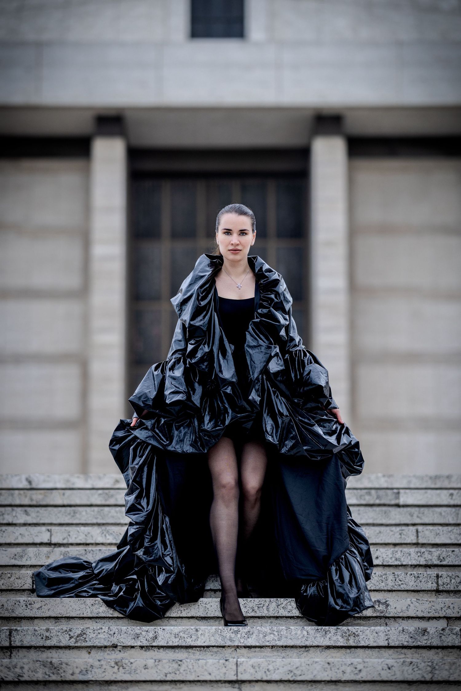 A model poses on stone steps in a dramatic black glossy ruffled gown with an avant-garde silhouette outside a neoclassical building.