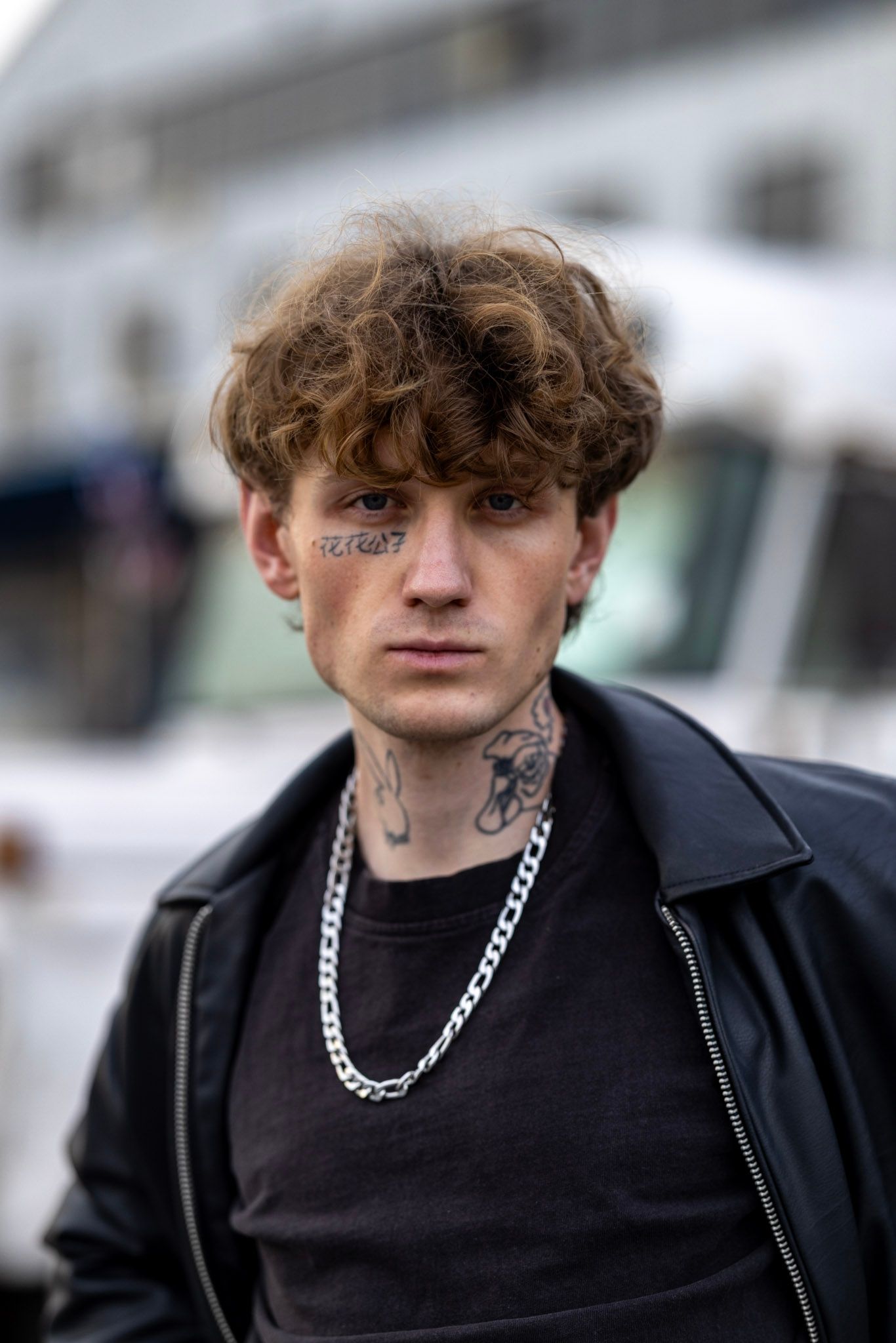 Young person with curly hair wearing black t-shirt, jacket and silver chain necklace poses outdoors with facial tattoos.