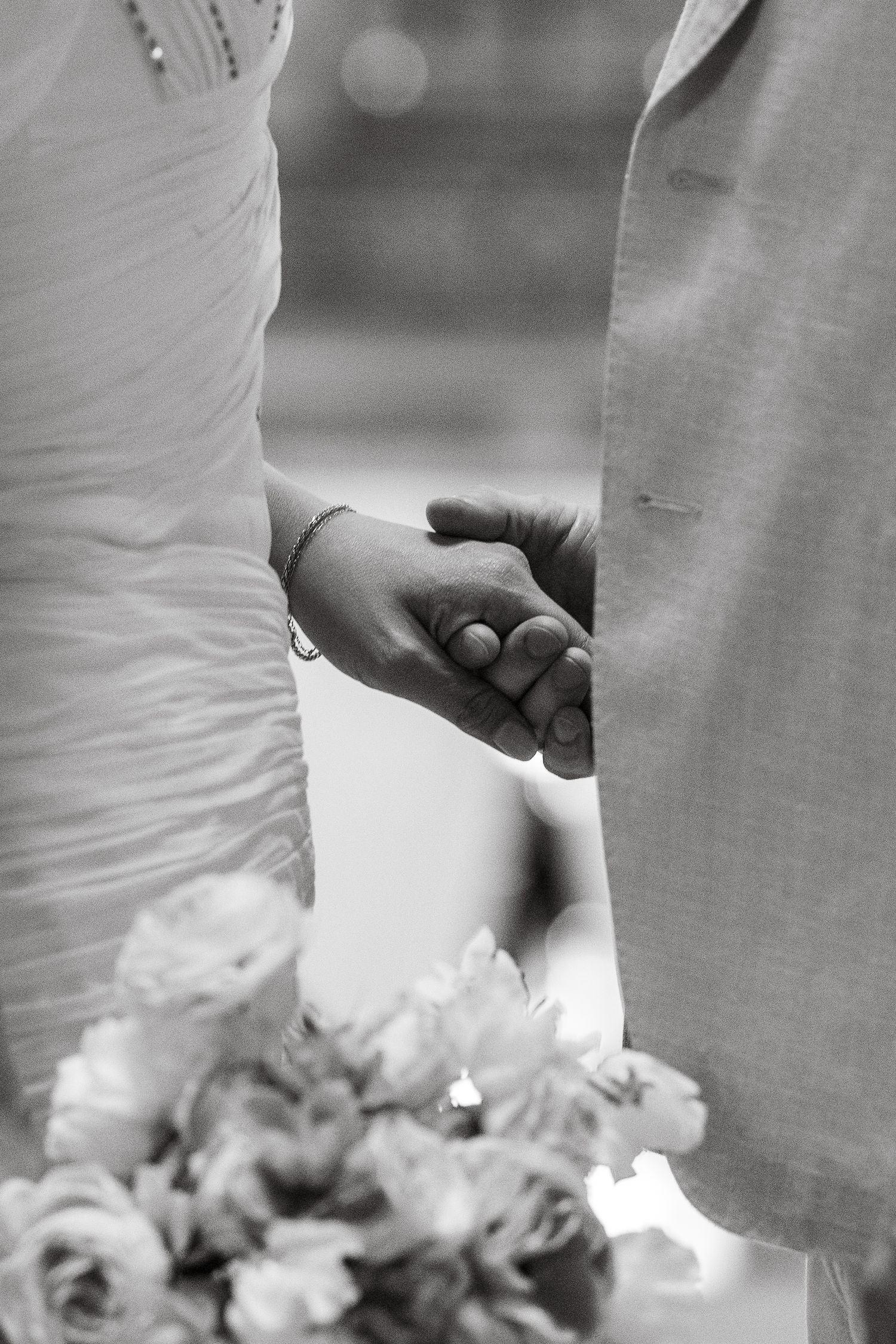 Black and white photo of bride and groom holding hands at wedding ceremony with bridal bouquet of roses.