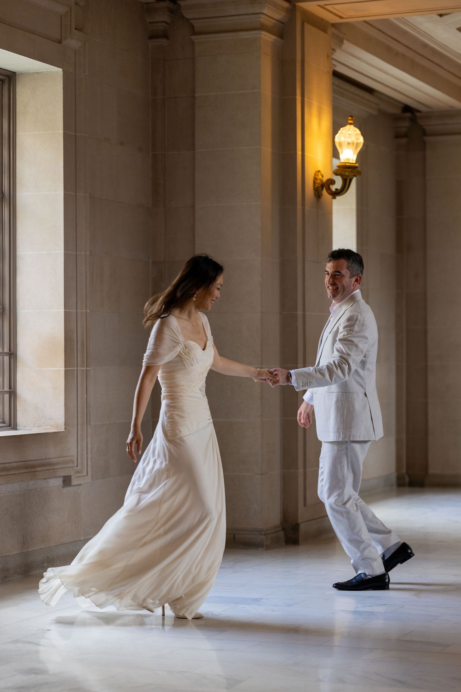 Bride and groom dancing in elegant stone hallway, woman in flowing white gown, man in white suit, warm wall sconce lighting.