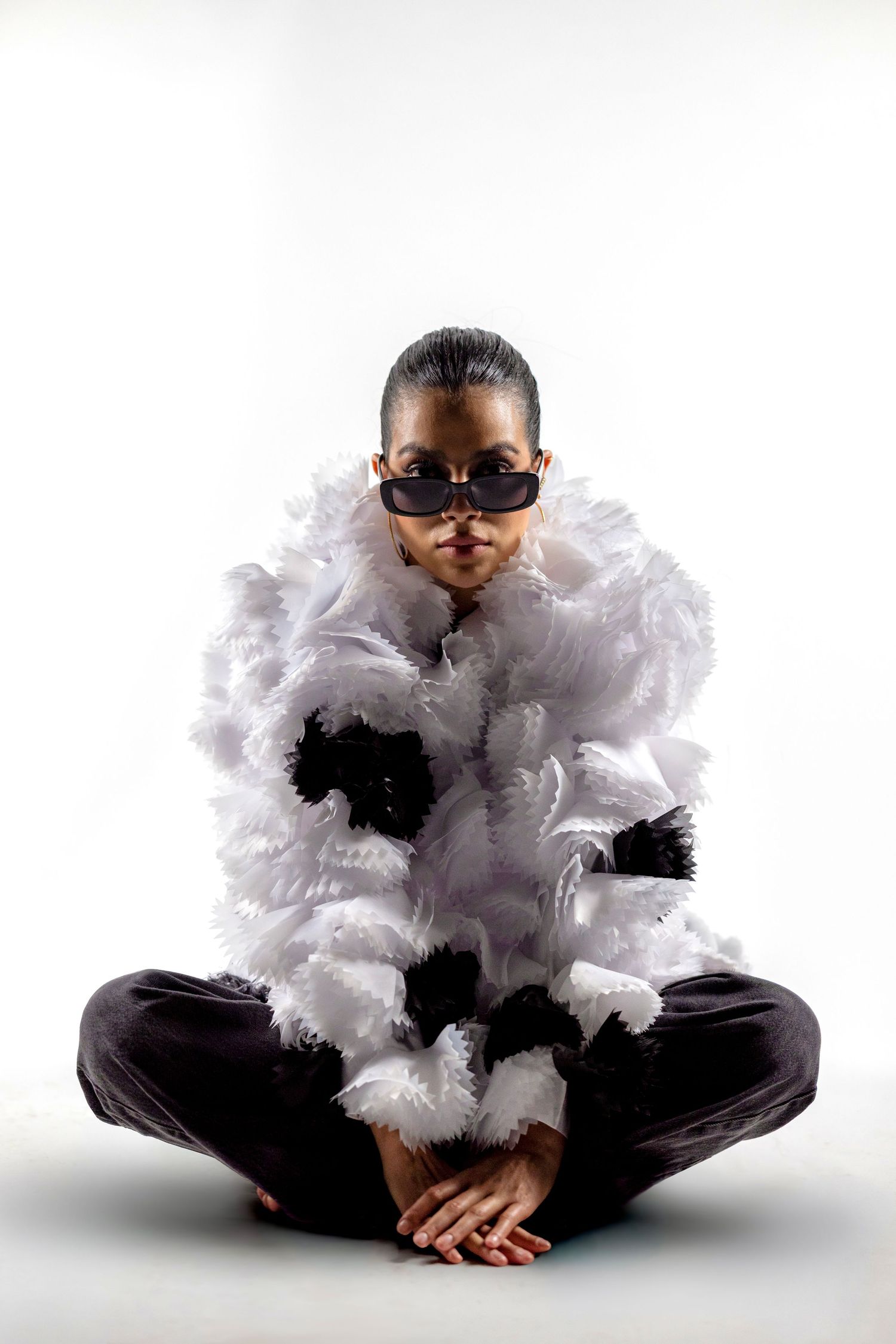 Person sitting cross-legged in a white feathered jacket with black accents and dark sunglasses on a white background.