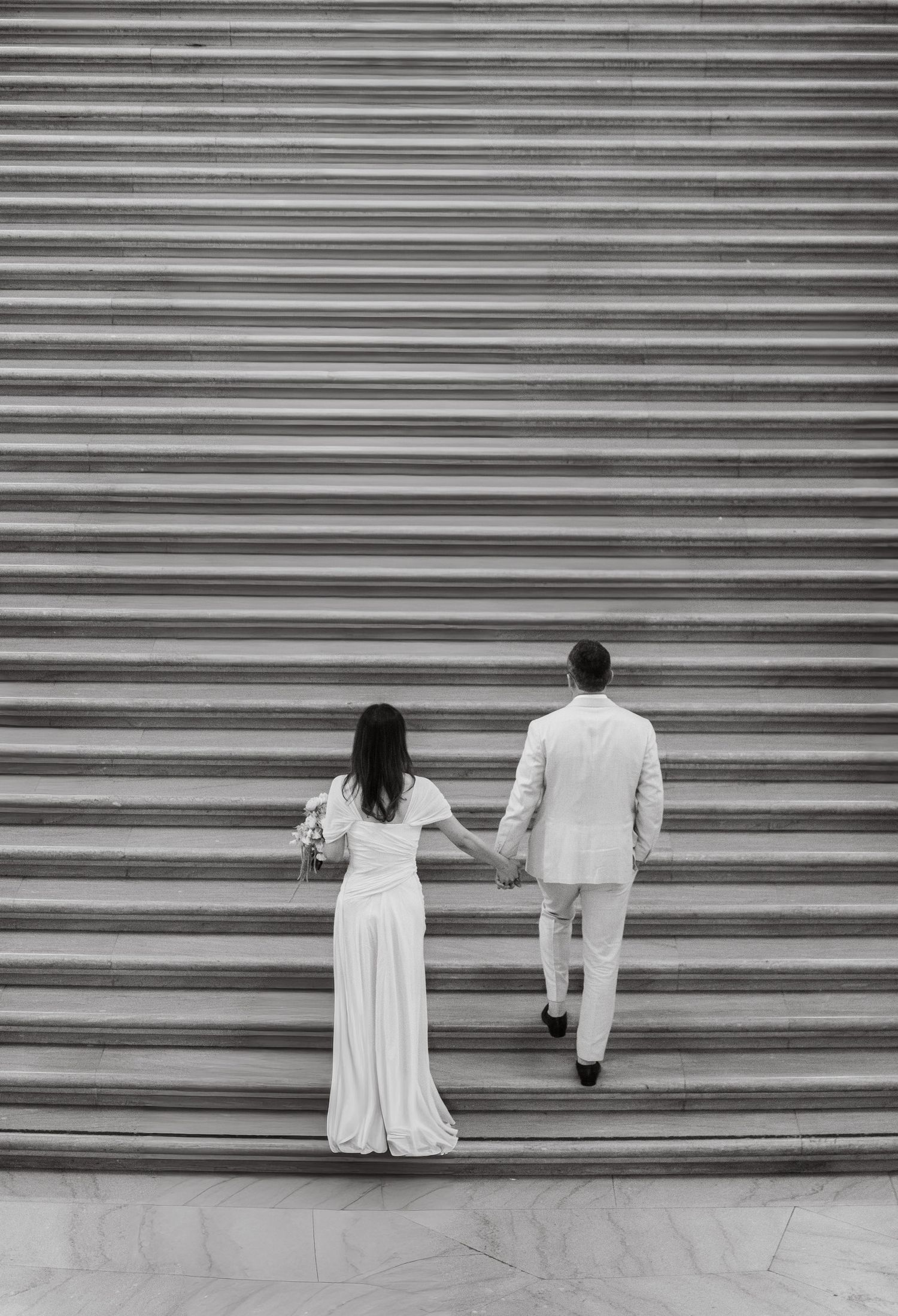 Black and white photo of a bride and groom holding hands walking up grand stone steps on their wedding day.