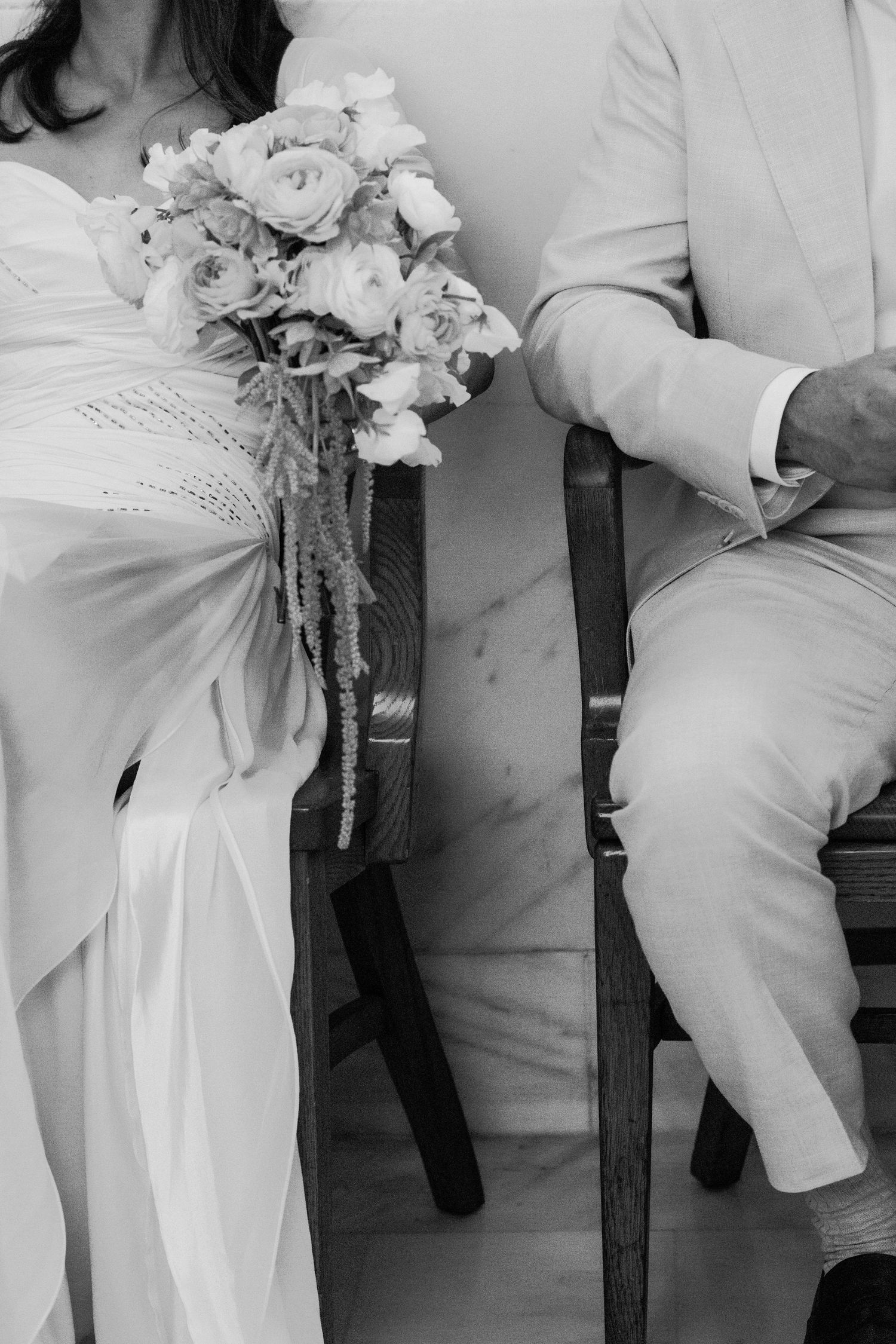 Black and white photo of bride and groom seated, bride holding elegant floral bouquet with roses and trailing greenery.