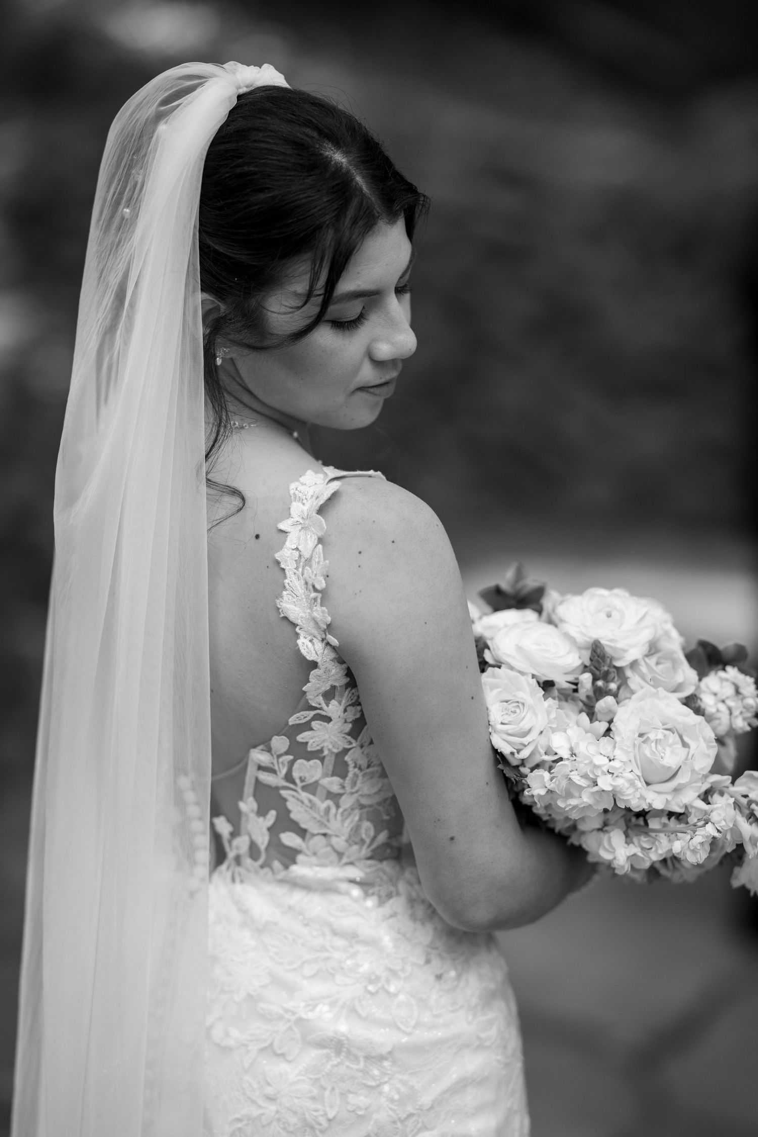Black and white portrait of a bride in lace gown and veil, holding a bouquet of roses, looking down gracefully.