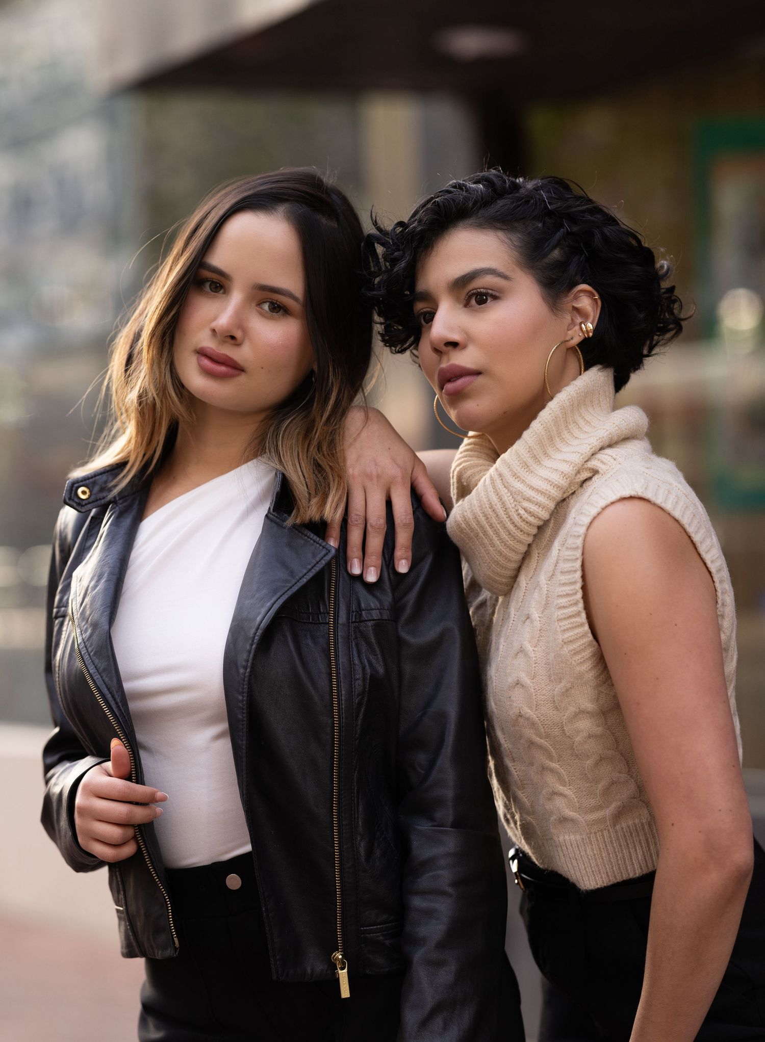 Two models pose together in contrasting black leather jacket and beige sweater outfits for a fashion photoshoot.