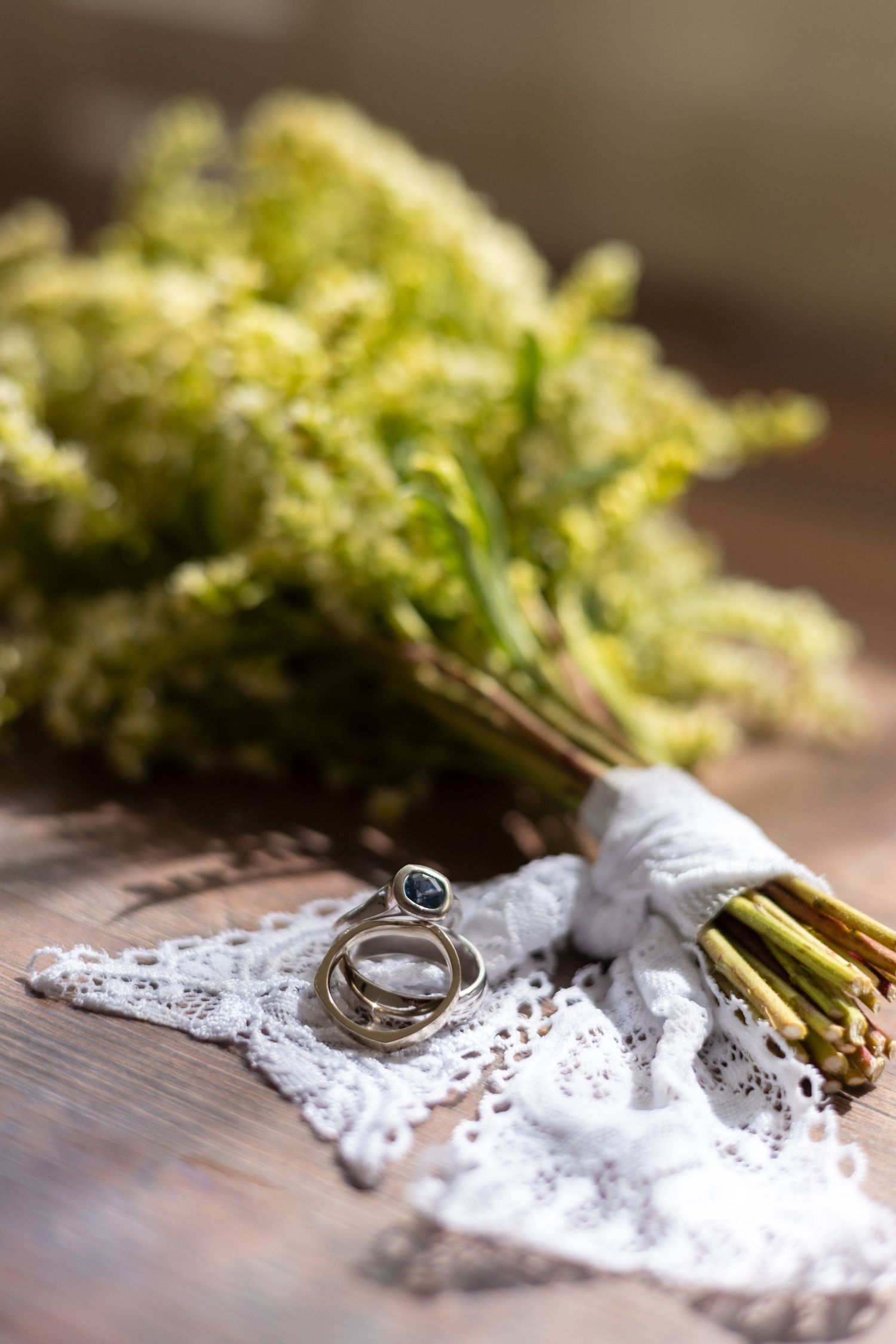 Wedding rings resting on lace fabric beside a rustic wildflower bouquet on a wooden surface.