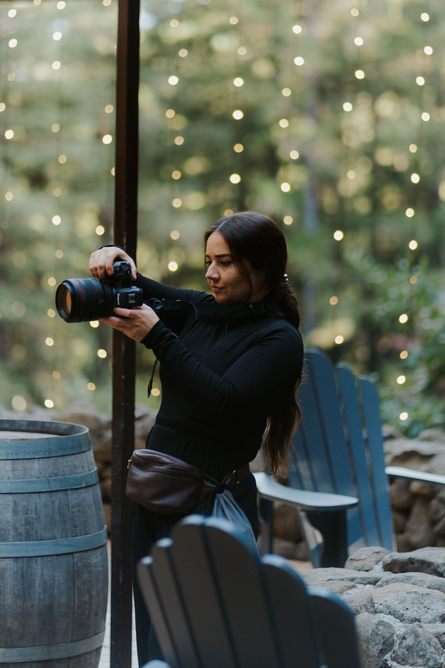 A photographer sits in an Adirondack chair taking photos with a camera against a backdrop of twinkling lights.