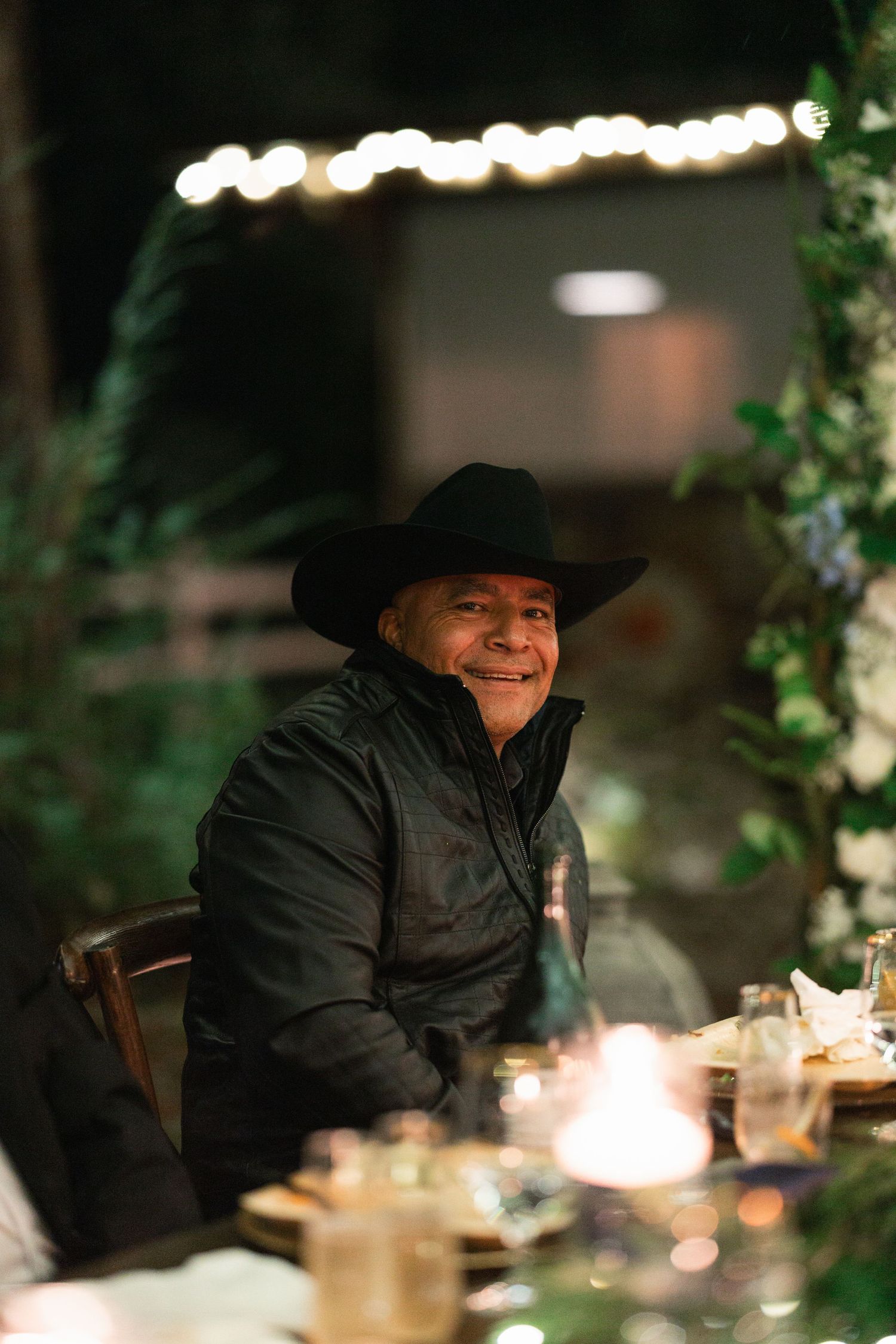 A person wearing a black cowboy hat and jacket sits at an outdoor dinner table illuminated by string lights.