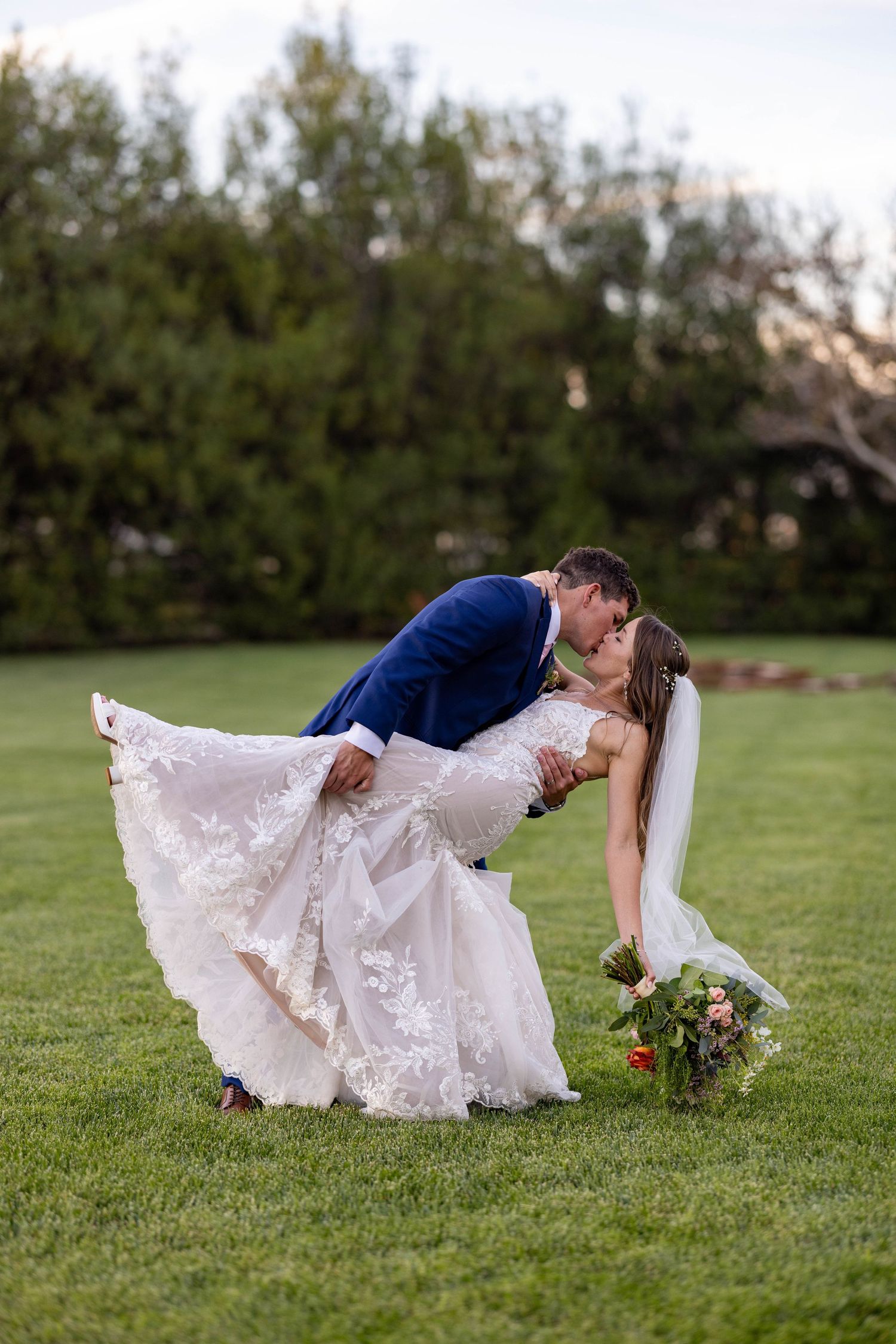 A bride and groom share a romantic dip pose on a grassy lawn during their wedding photoshoot at sunset.