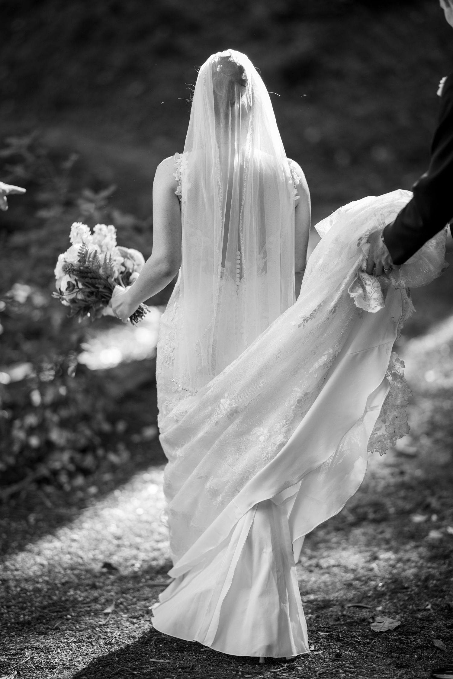 Black and white photo of a bride walking away, wearing a flowing gown and veil, holding a bouquet.