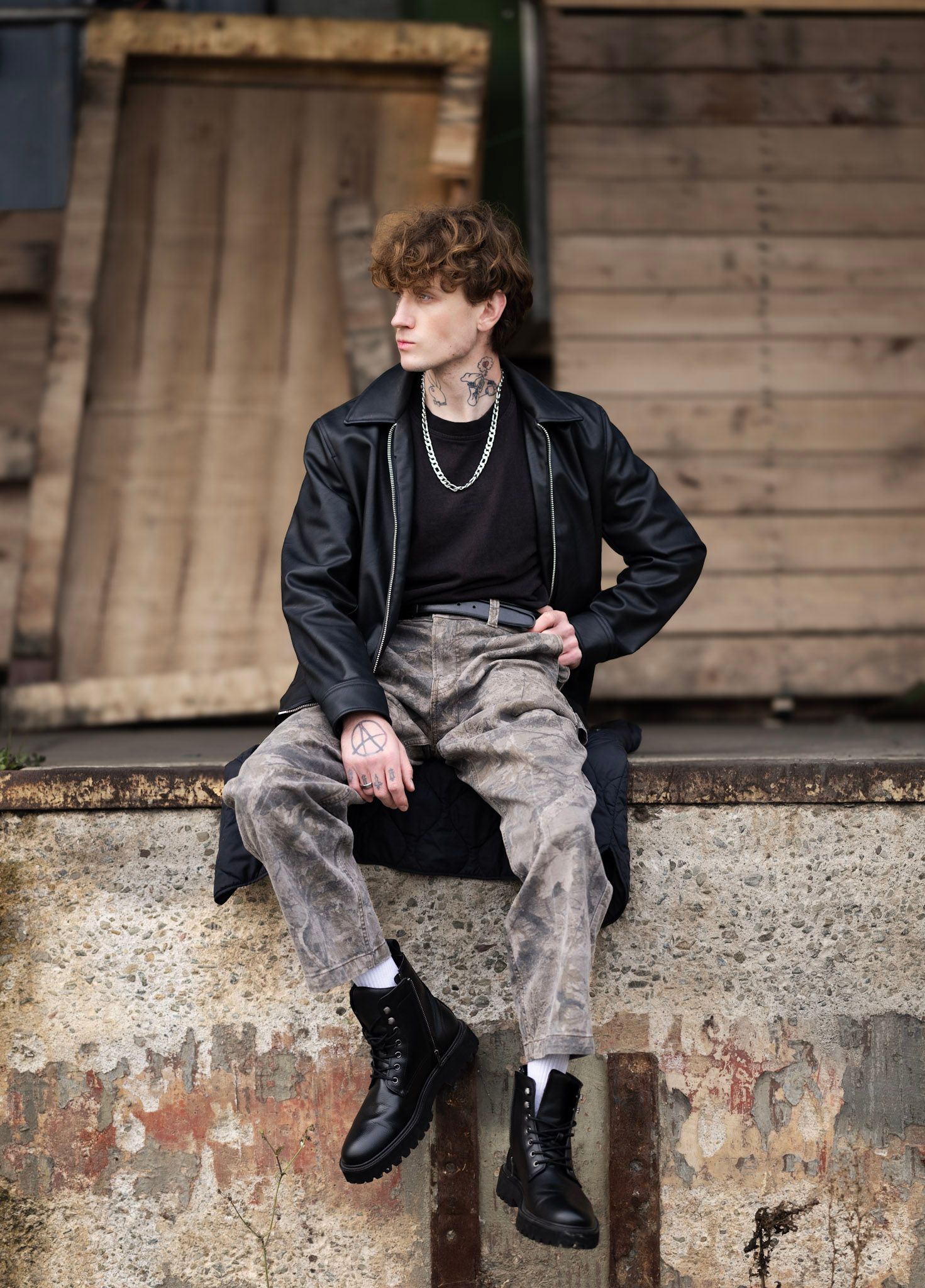 A person in a black leather jacket and distressed jeans sits on a concrete ledge wearing combat boots and a silver chain.
