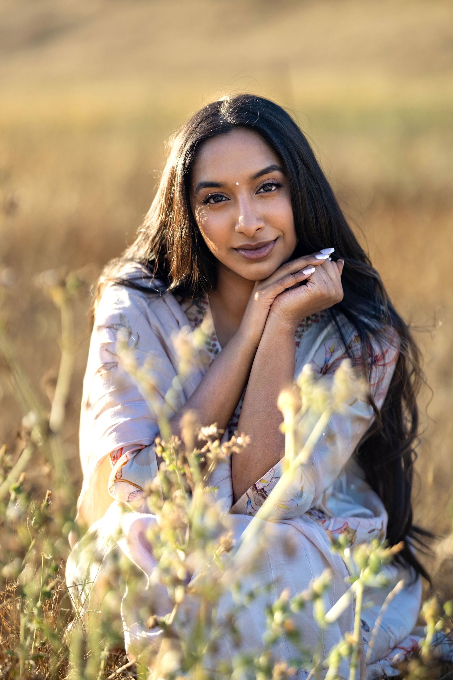 Young South Asian woman with long dark hair sits in golden field wearing floral dress, hands clasped near chin.