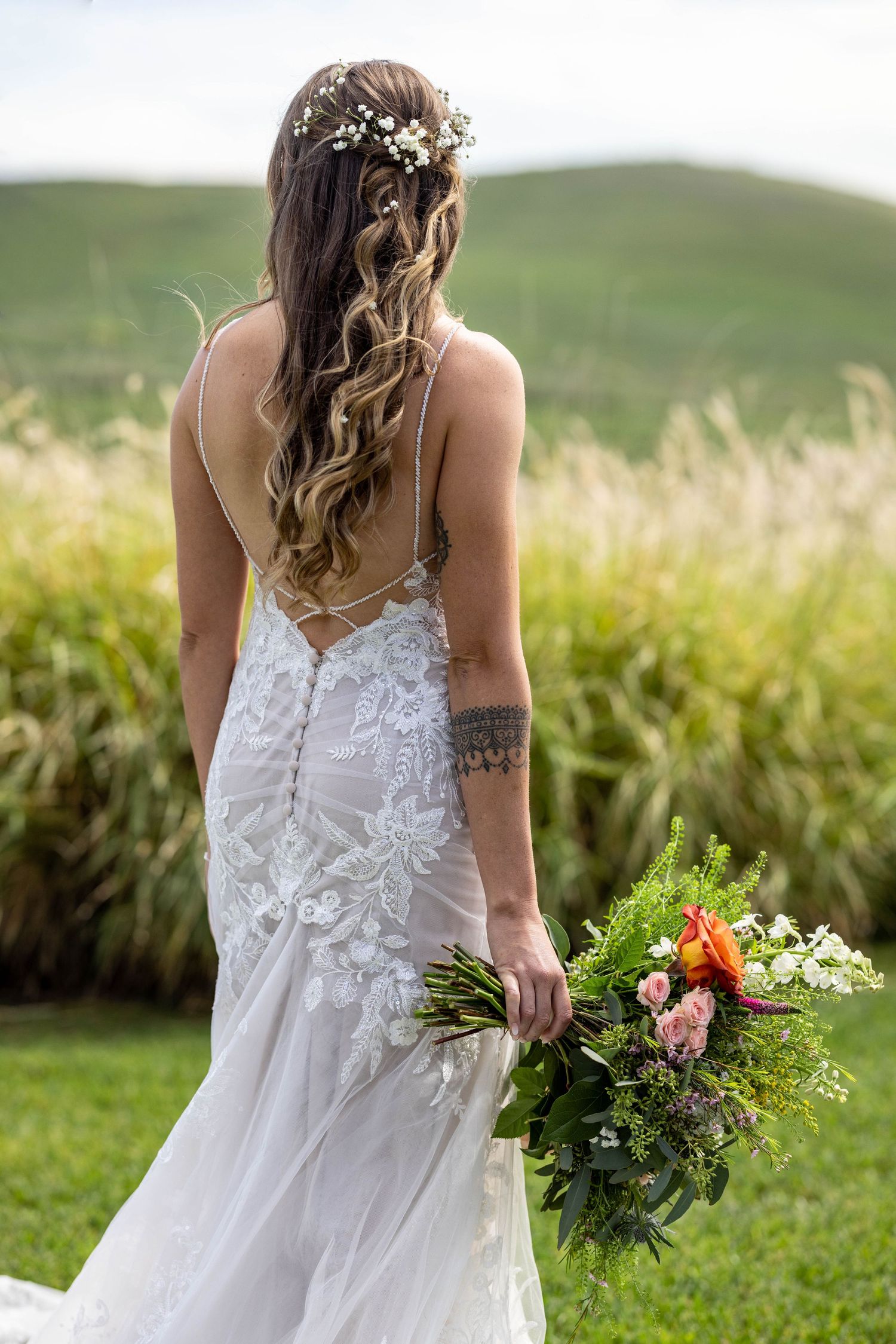 A bride in a white lace wedding dress holds a colorful wildflower bouquet while standing in a sunlit meadow.