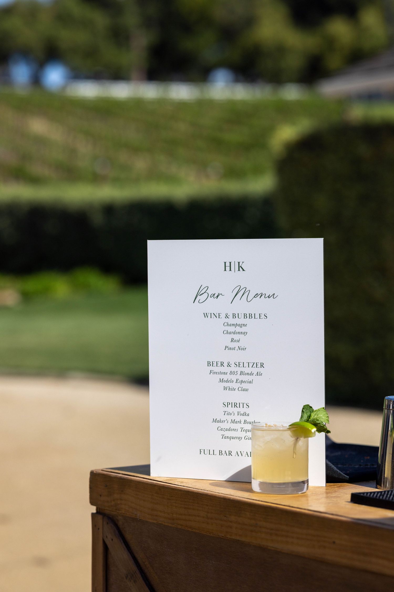 A cocktail menu and refreshing drink with lime garnish sit on a wooden surface against a blurred outdoor background.