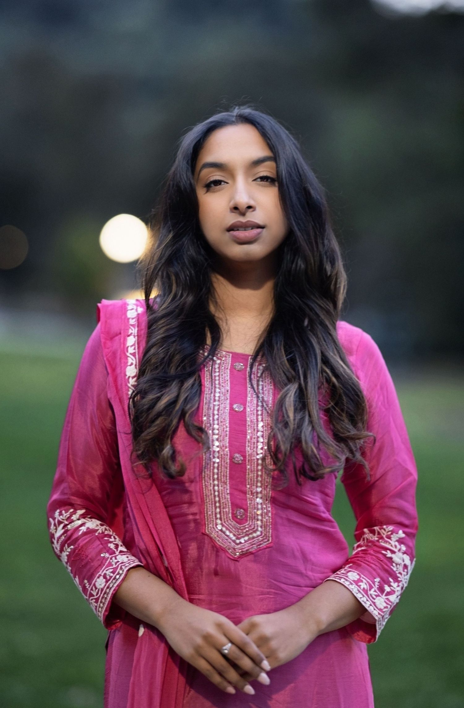A person in a bright pink embroidered traditional tunic poses outdoors at dusk.