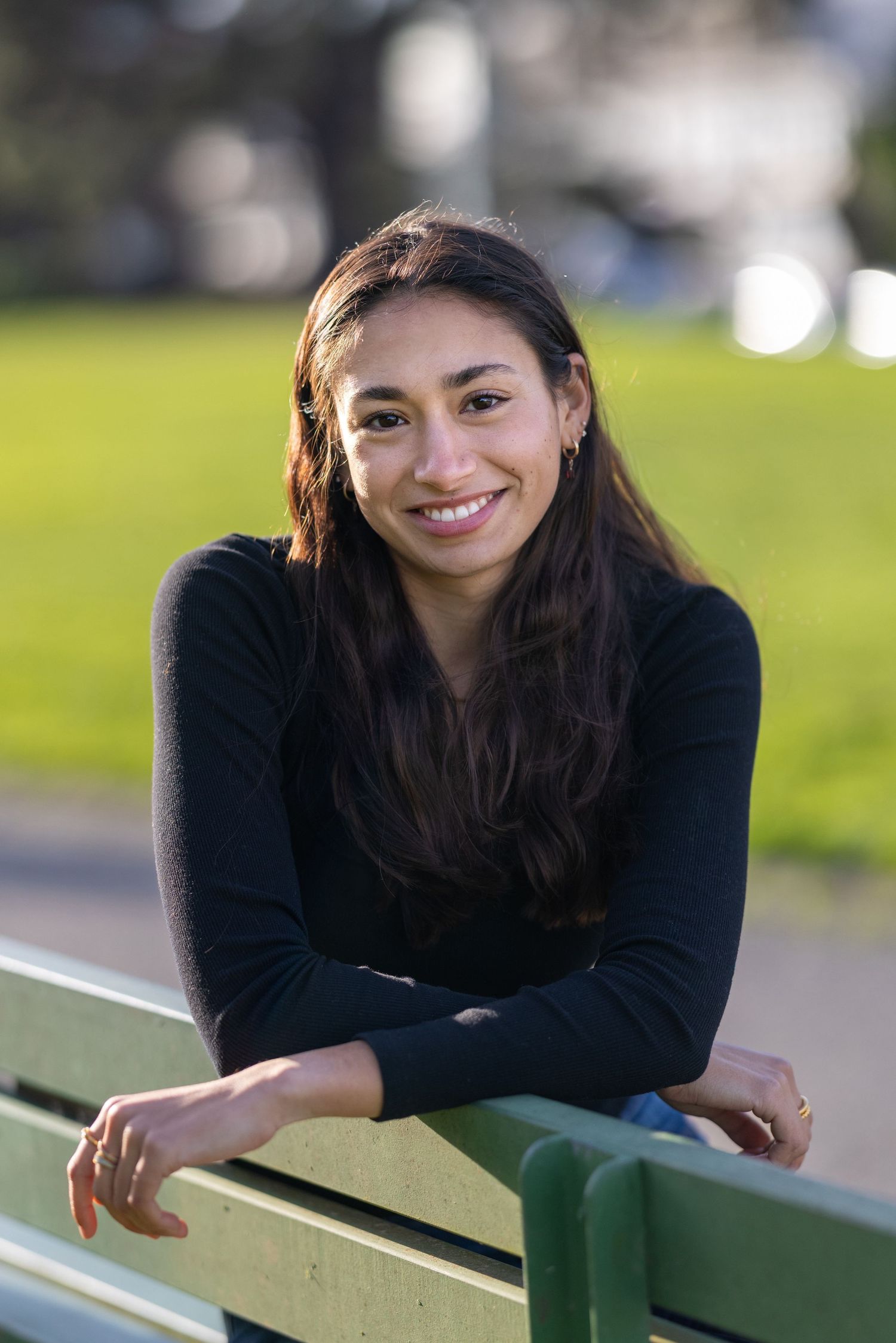 A young woman in a black long-sleeve shirt smiles while leaning on a railing outdoors on a sunny day.