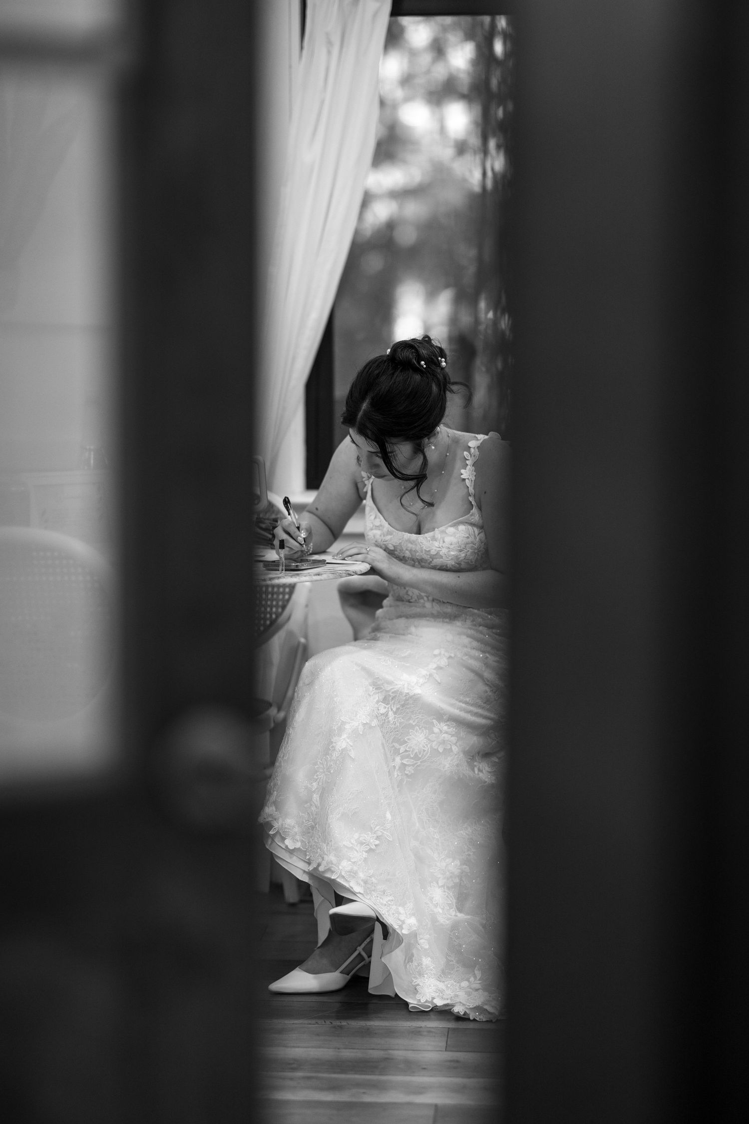Black and white photo of a bride in a lace gown sitting and writing, viewed through a doorway with curtains.