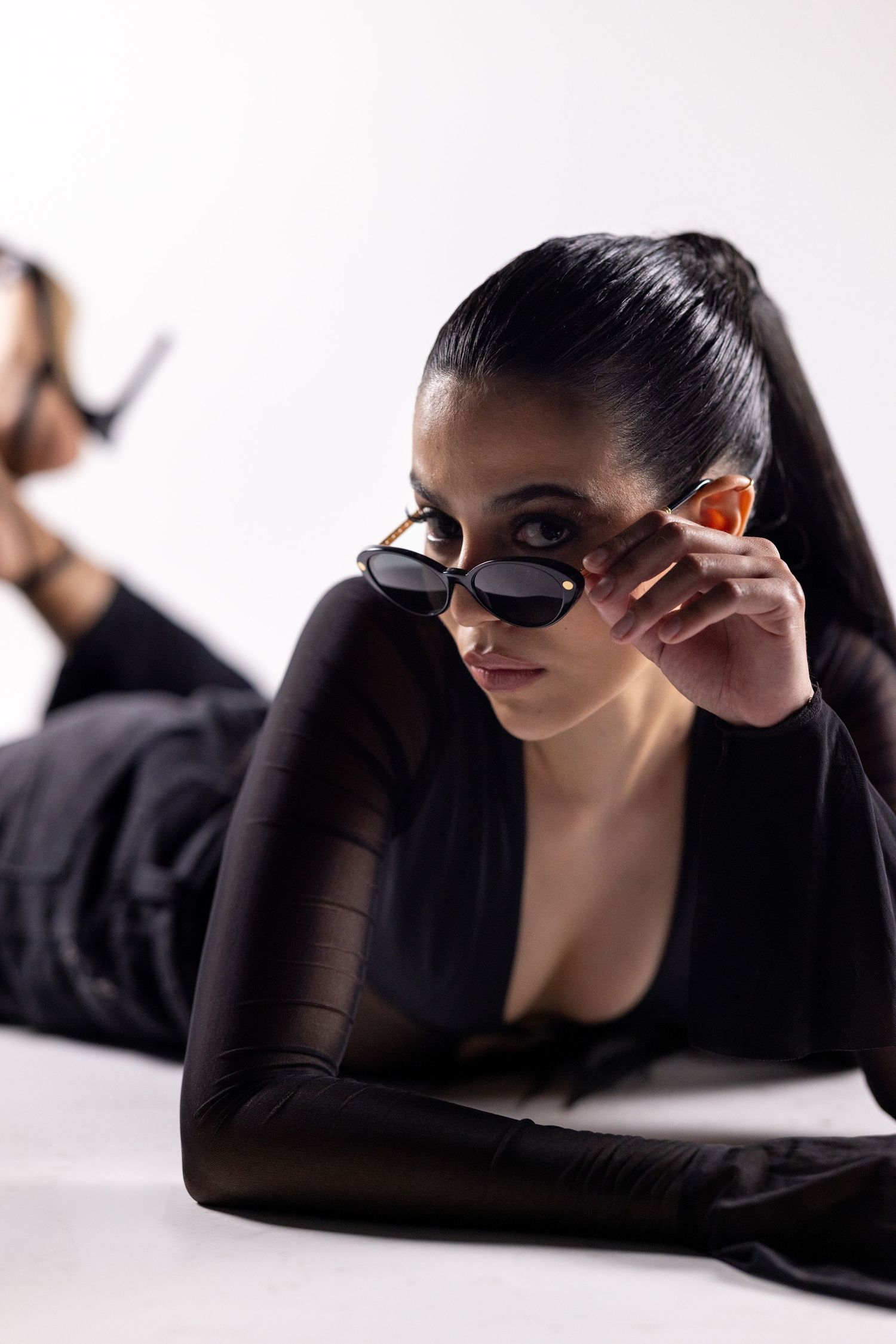 Woman in black outfit lying down, adjusting stylish cat-eye sunglasses, with sleek ponytail on white background.