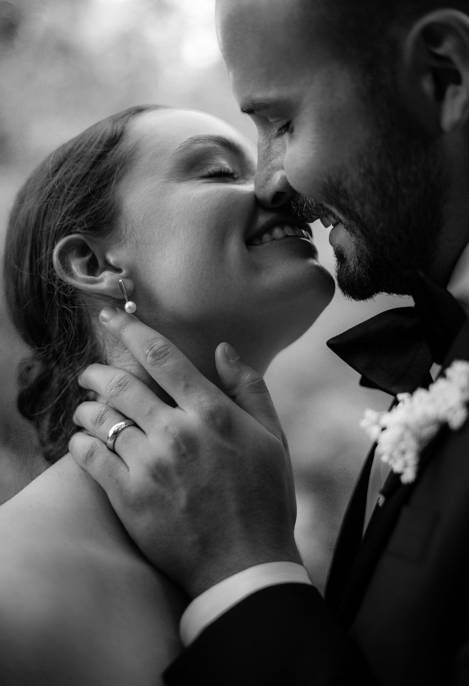 Black and white photo of a smiling bride and groom sharing a romantic moment, with wedding rings and boutonniere visible.