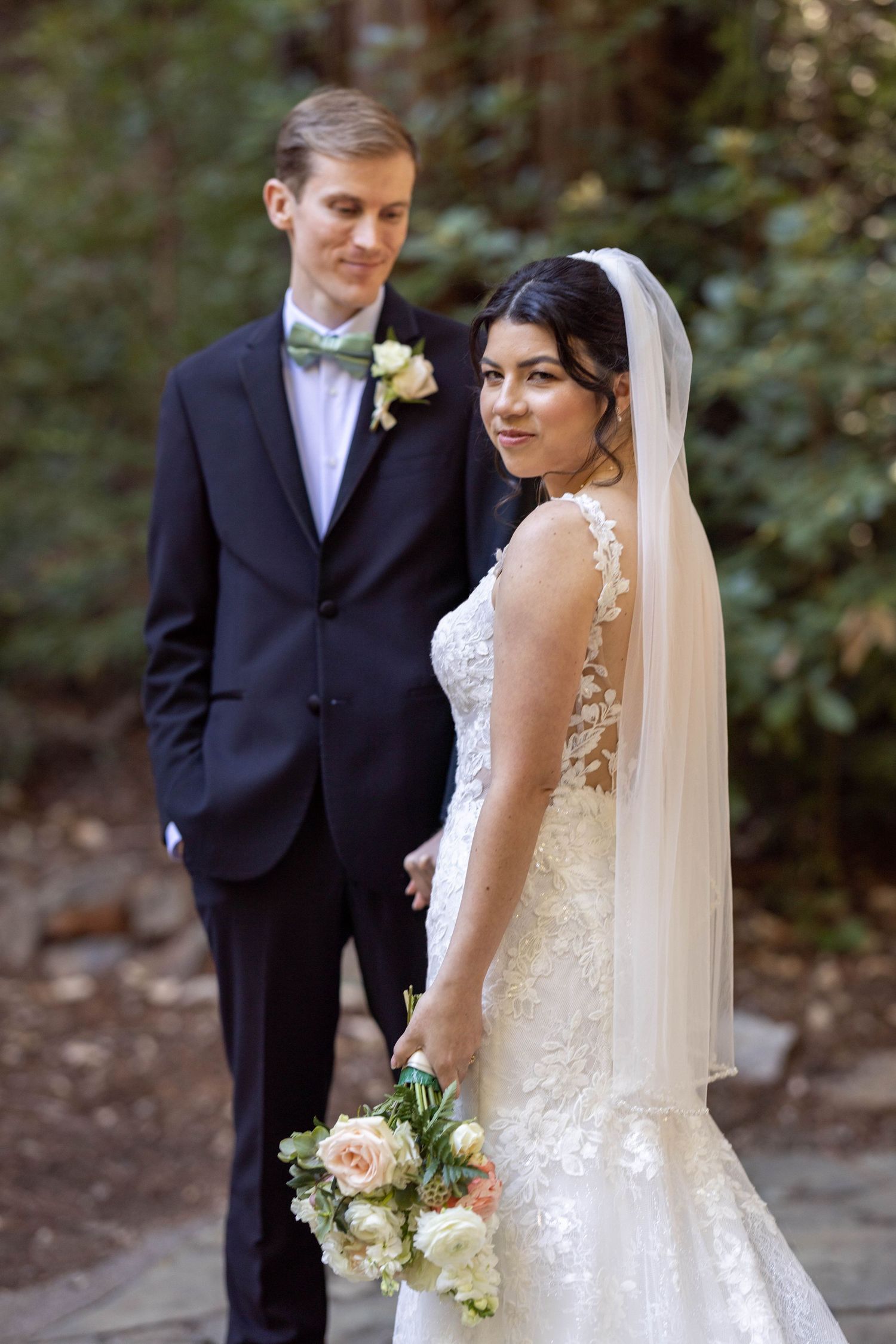 Bride in lace gown holding bouquet looks back at camera while groom in navy suit stands behind her outdoors.
