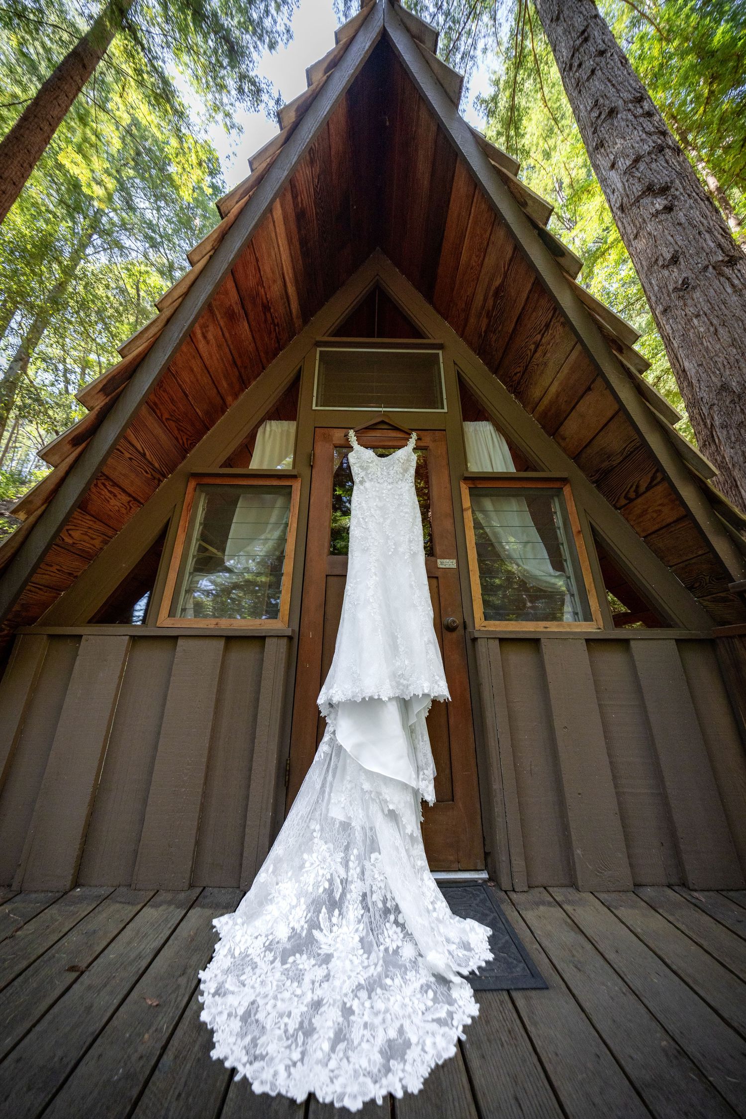 White lace wedding dress with long train hanging outside an A-frame cabin surrounded by tall trees.