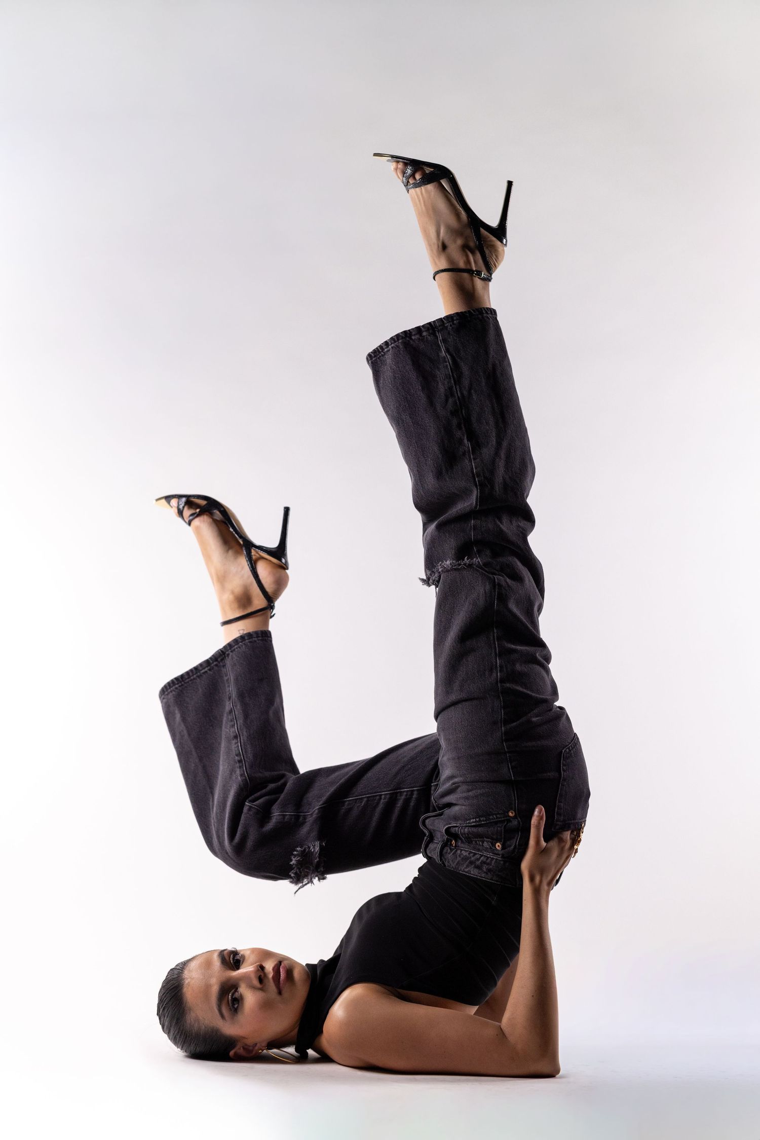 Woman in black jeans and heels performing a shoulder stand pose against a white background.
