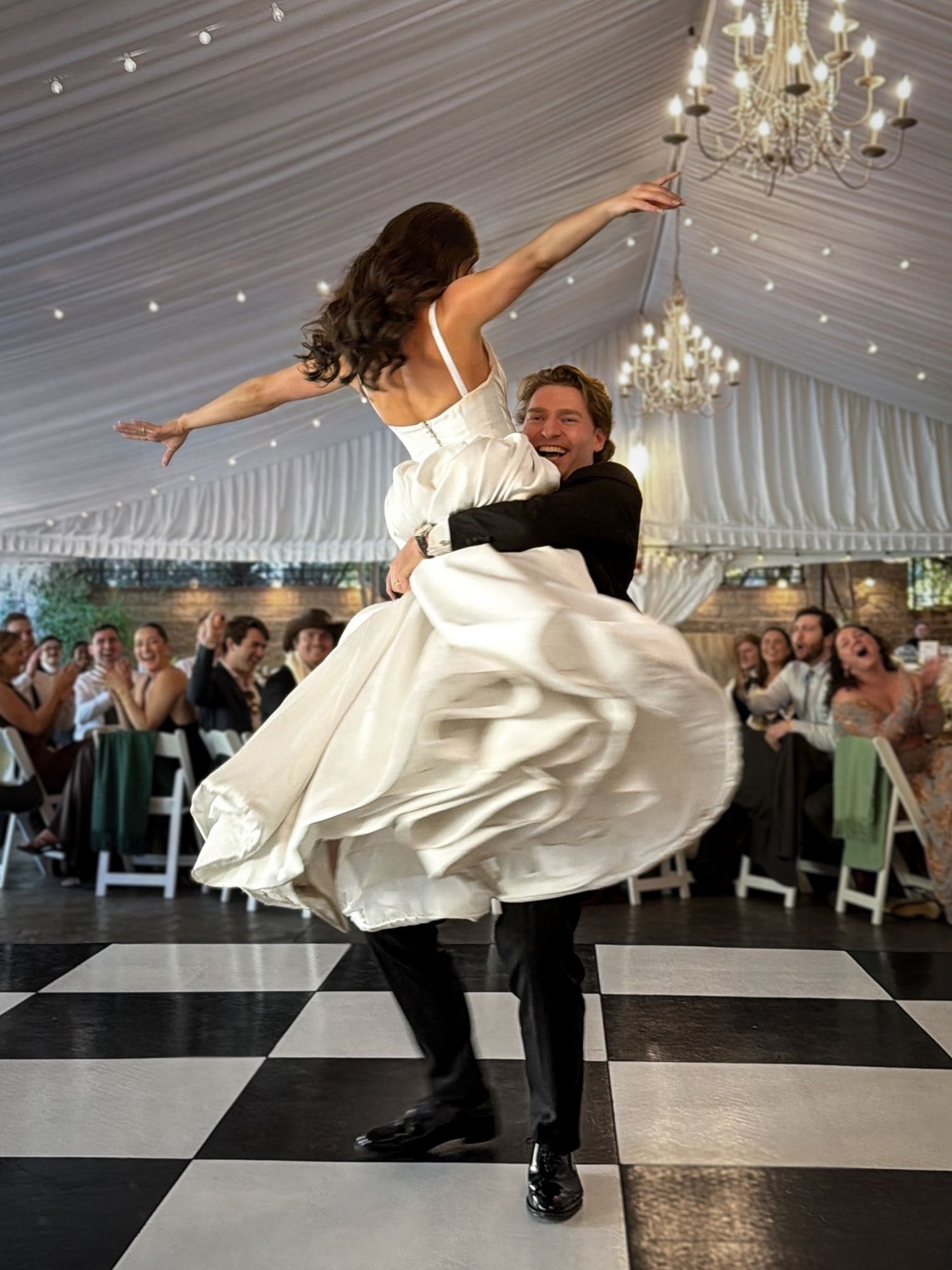 A couple performs a dramatic dance lift on a black and white checkered floor under elegant chandeliers at their wedding reception.