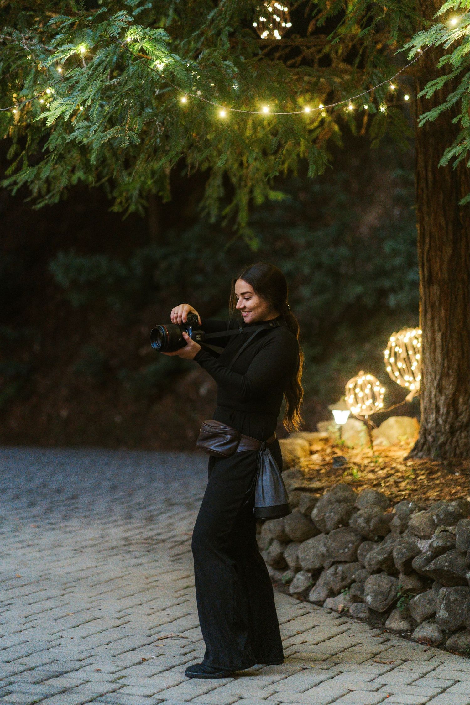 A photographer in black clothing takes photos in a garden setting with string lights and stone walls at dusk.