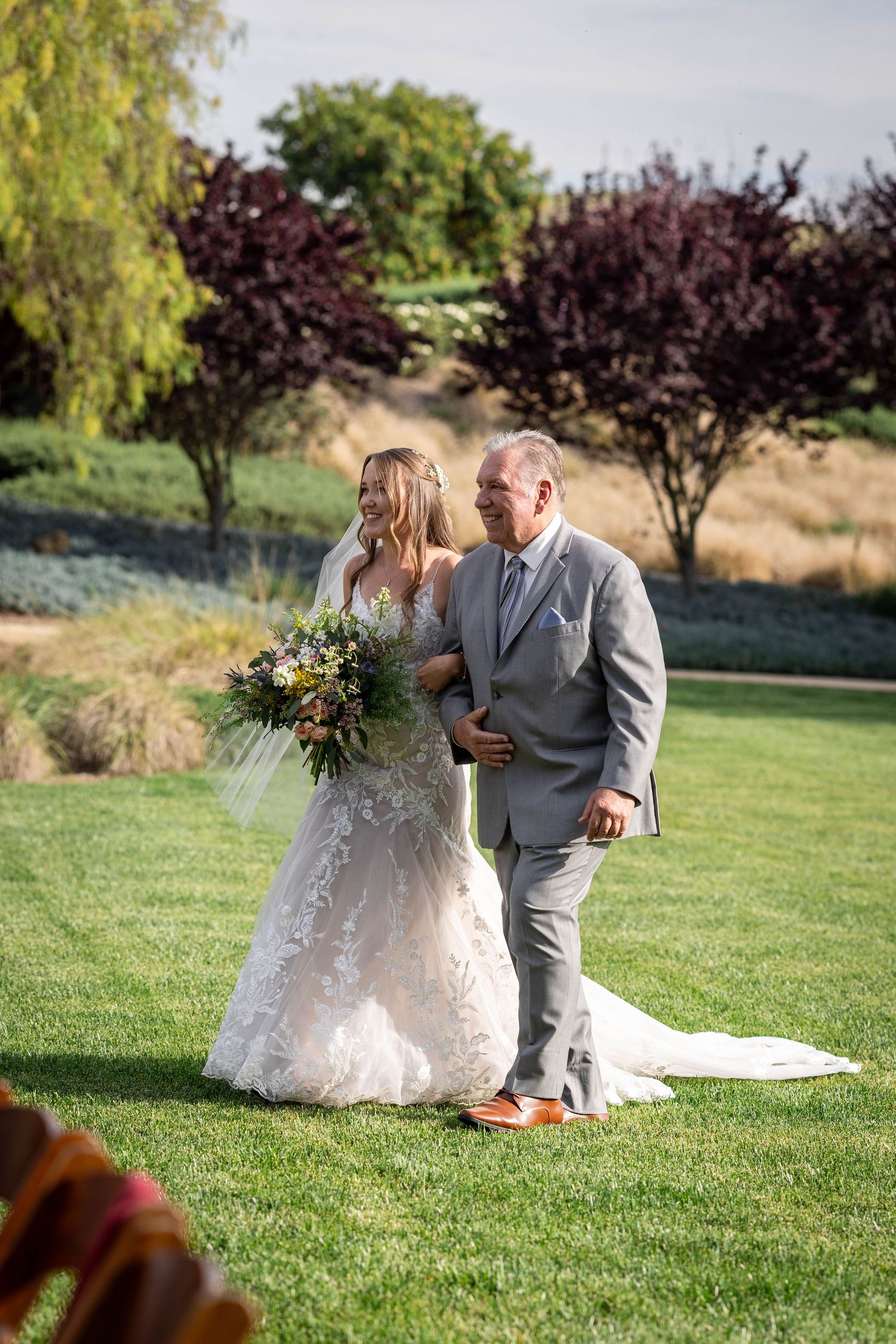 Bride in lace gown walks with older man in gray suit across green lawn, holding wildflower bouquet at outdoor wedding.