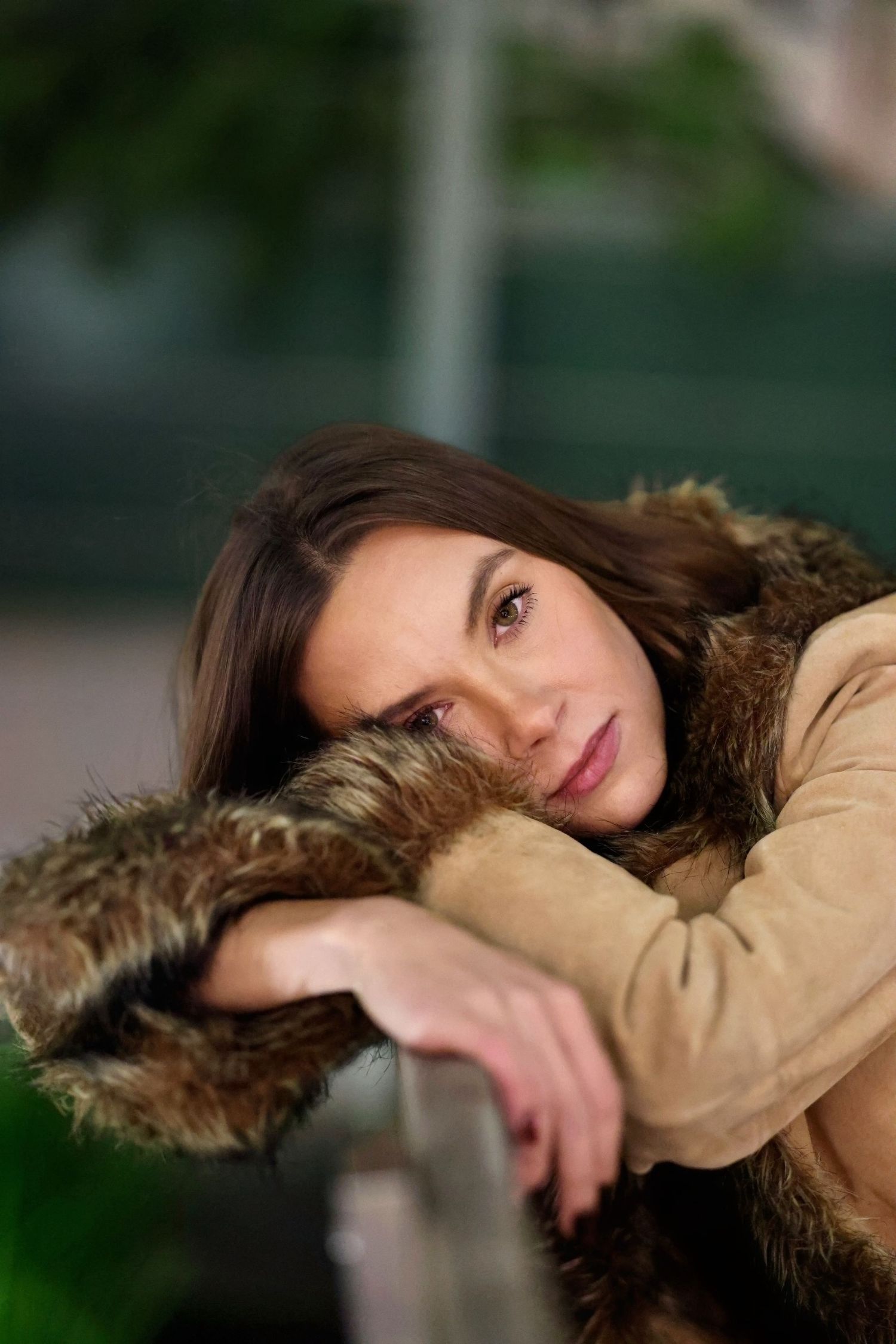 A woman in a tan fur-trimmed coat poses against a blurred green outdoor background.