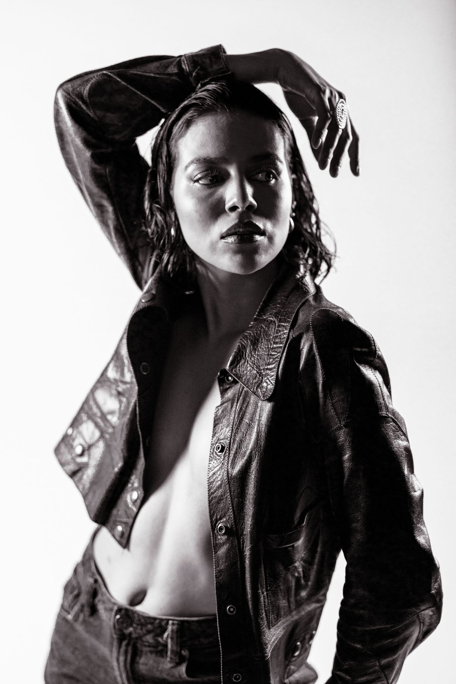 Black and white portrait of a woman in an open leather jacket posing with arm raised against a light background.