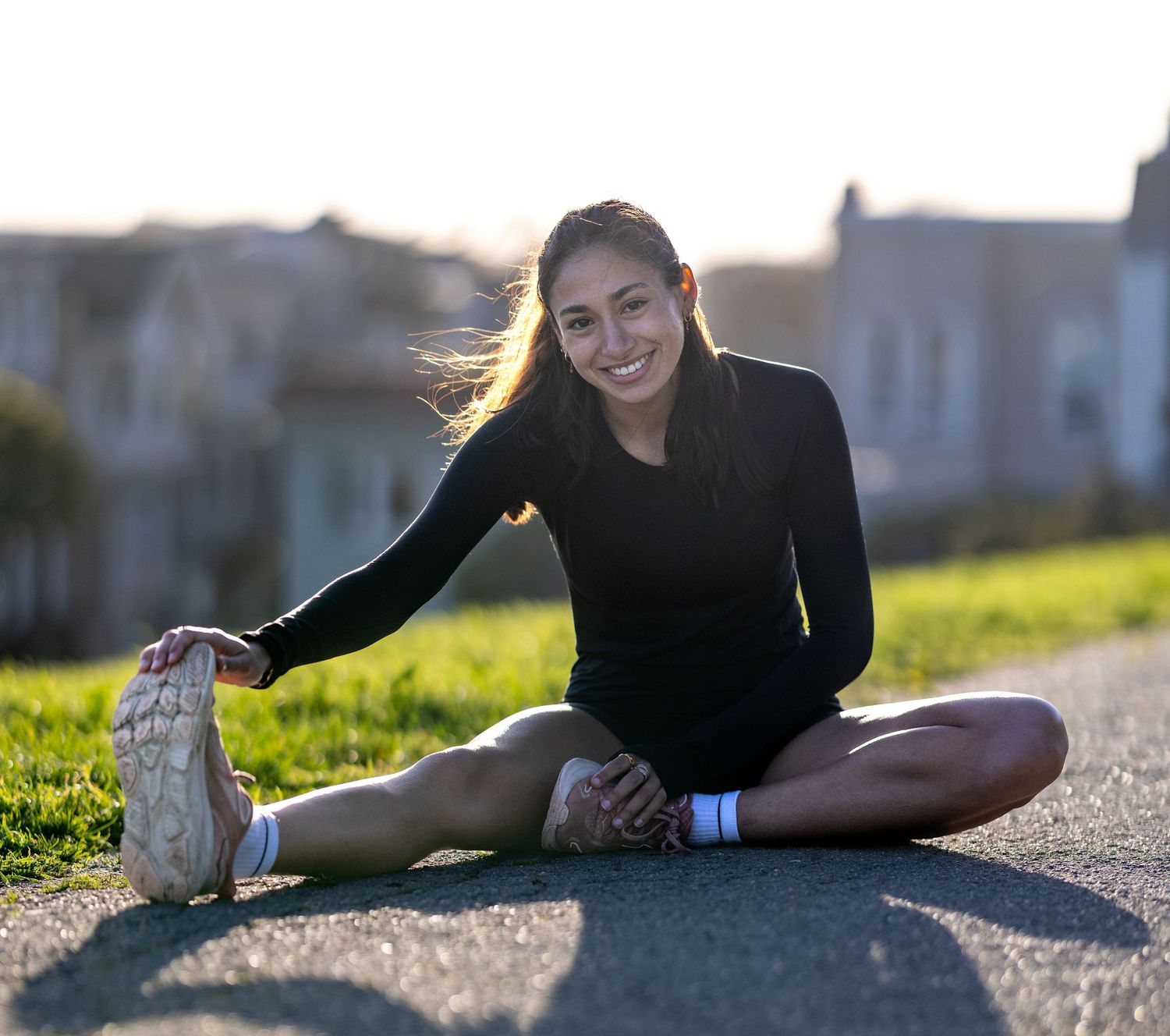 Runner stretches on path during golden hour with sunlit grass and buildings in background.